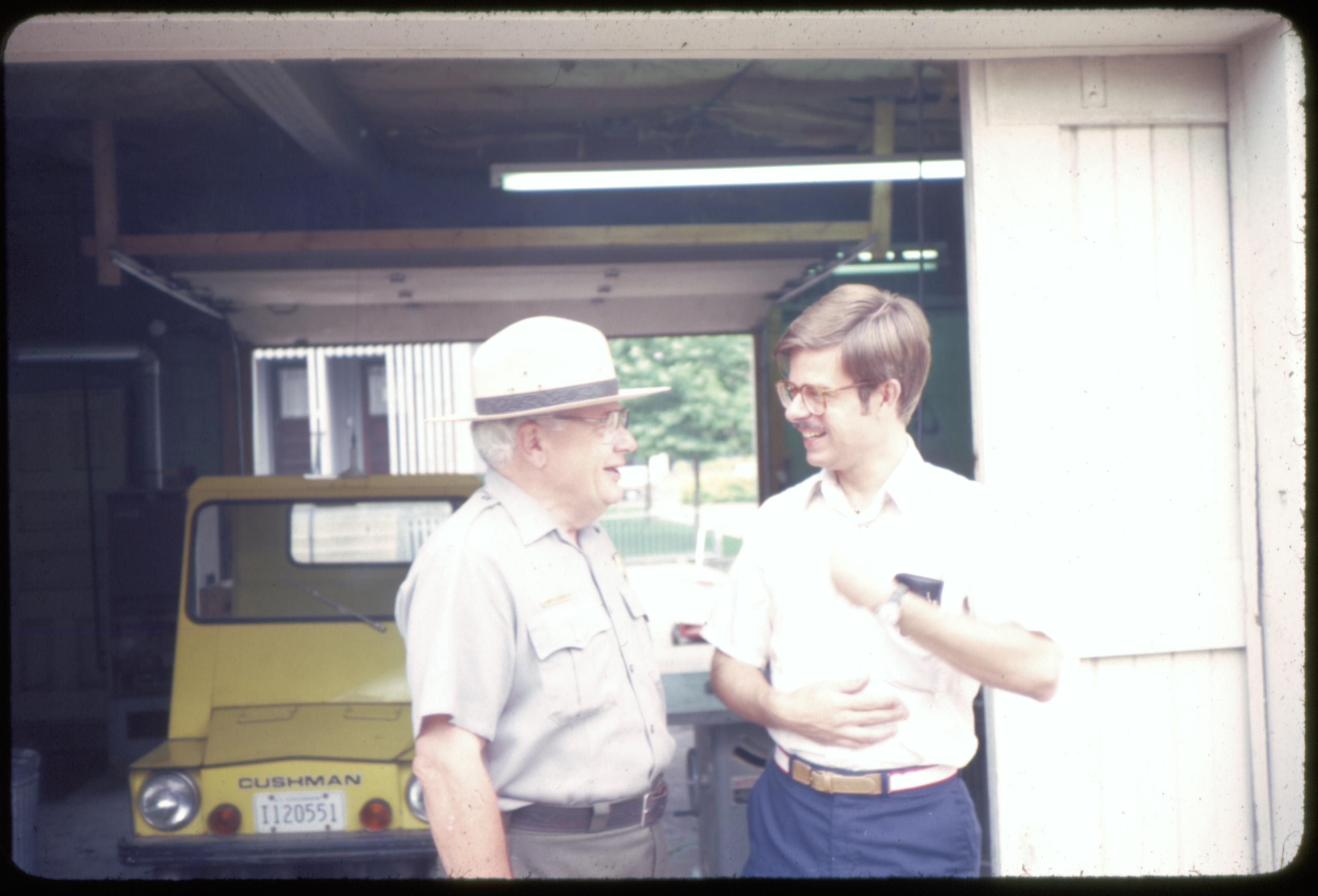 Supt. Al Banton talking to gentleman in the alley by the Maintenance Shop.  Stuve House west porch visible in background. Looking west staff, Maintenance Shop, Stuve, Cushman