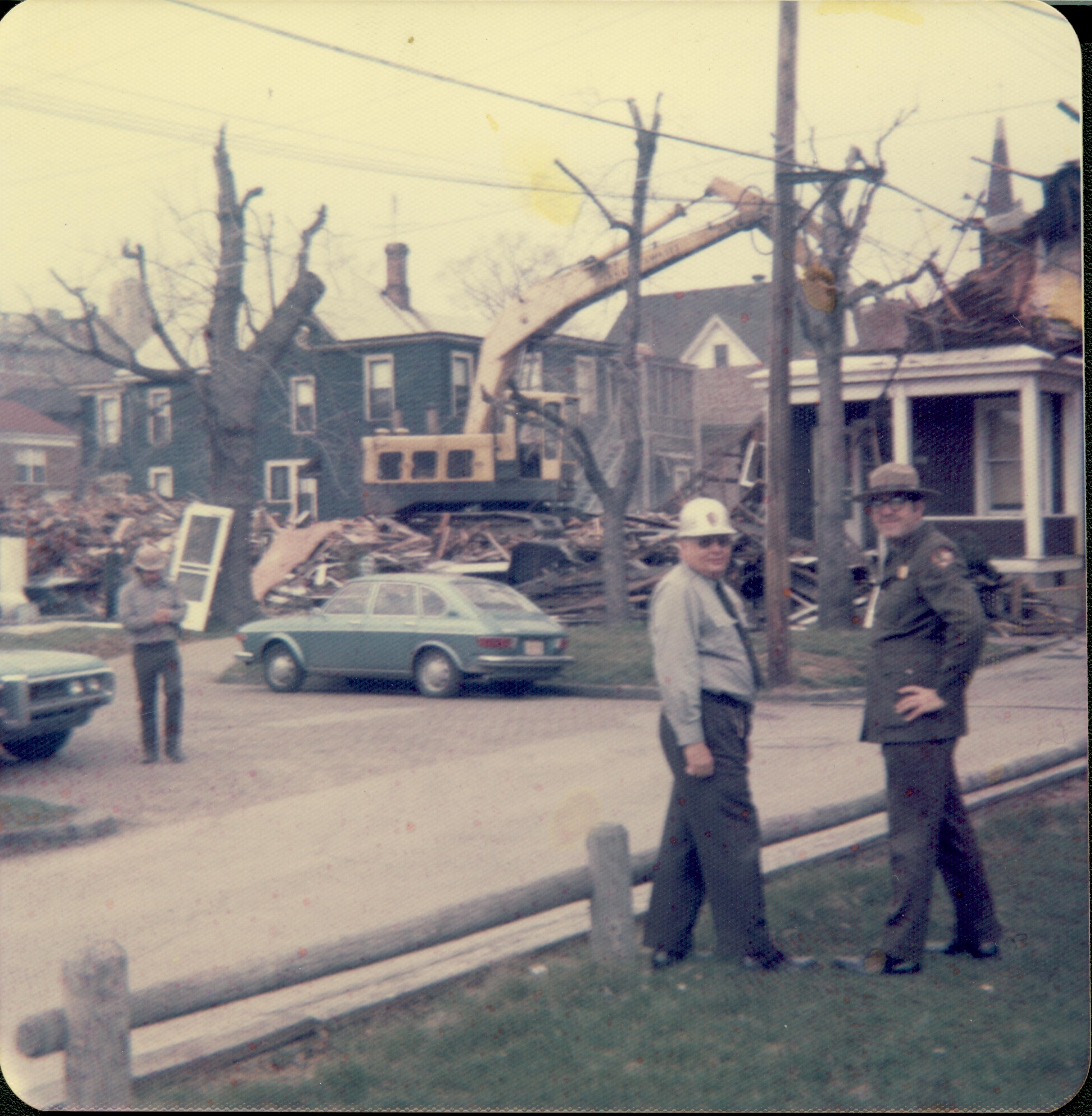 House owned by William Hermes in process of being razed, Block 7, Lot 8, along 8th Street.  Dark house in background owned by Eleanor Pruet. Maintenance Worker Tom Pacha in hard hat on left, Supt. Al Banton in hard hat center, and Ranger Chet Hamilton watch progress. Area is now Visitor Center. Looking Northwest from South of Jackson Street along alley. Hermes, Pruett, Jackson Street, razing, Visitor Center