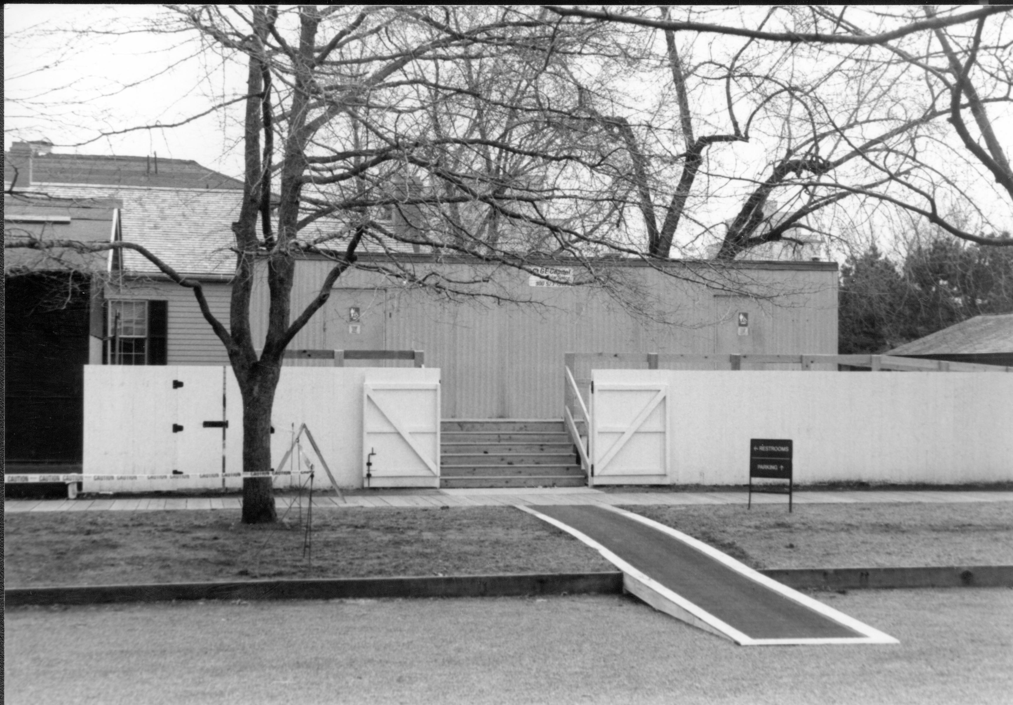 Temporary restroom trailer in use while Visitor Center was remodeled. Located in the Corneau lot (Block 6, Lot 16). Wheelchair access ramp goes off decking to right, steps come down front to another access ramp into Jackson Street. Corneau House (under restoration) on far left. Sprigg House in background left. Maintenance shed on background far right. Looking South from boardwalk on North side of Jackson Street. Visitor Center, Corneau, restroom, trailer, Jackson Street, shed