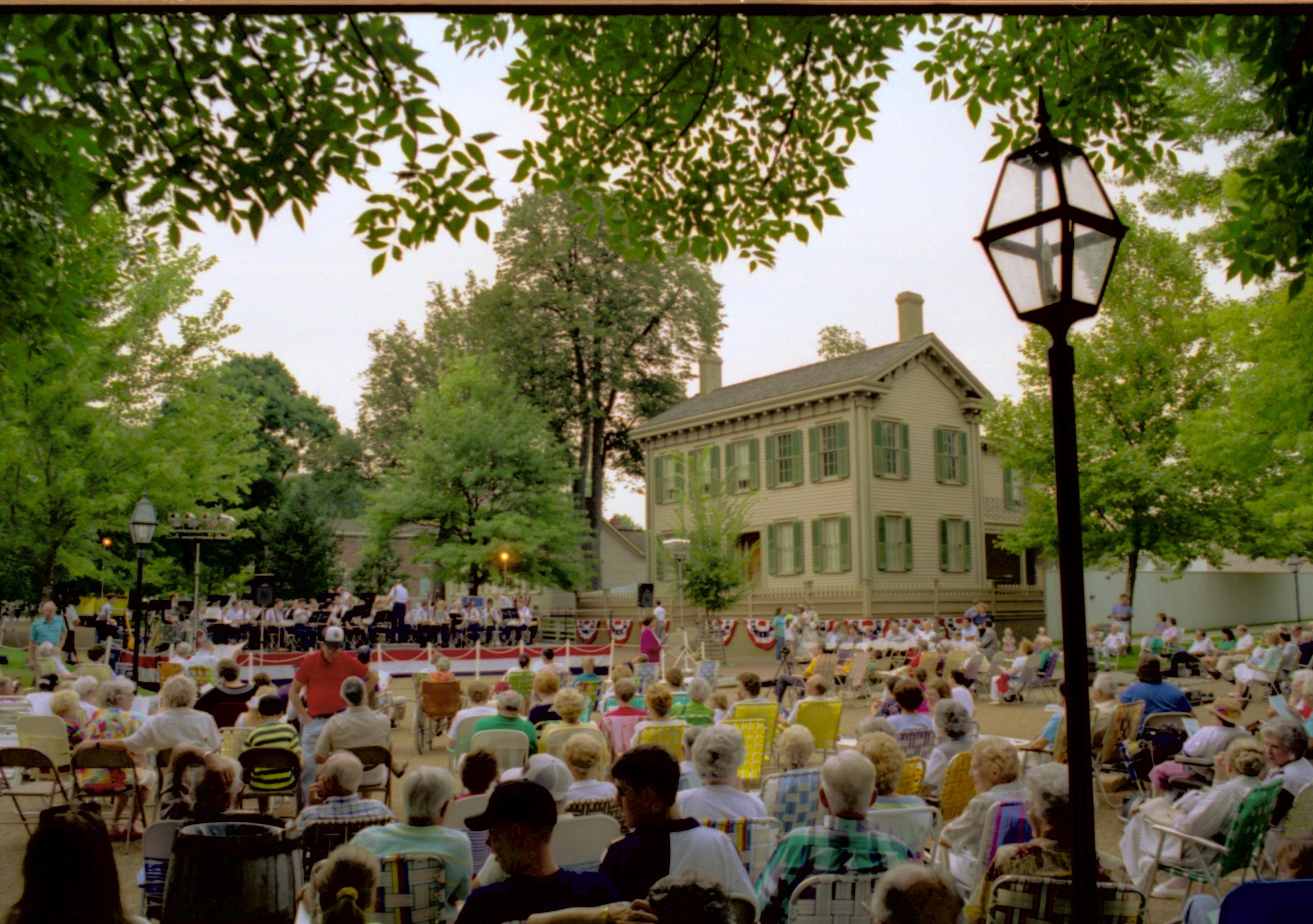 Public seated, band on platform and Lincoln home in background. Lincoln Home NHS- Coast Guard Band Concert, 267618 concert, Coast Guard