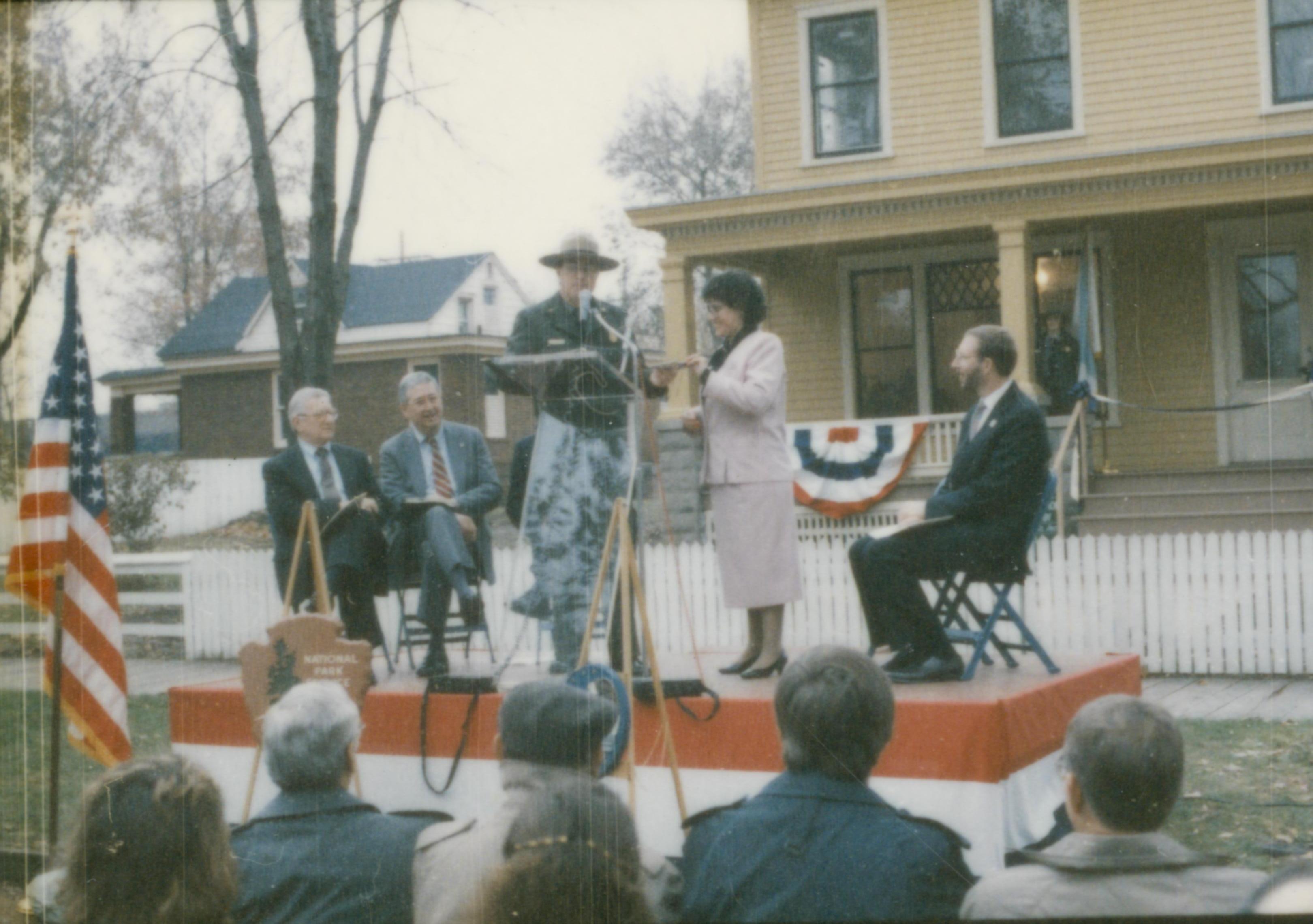 Five people on speakers platform in front of Cook house. Lincoln Home NHS- Cook House Re-opening, 1 Cook, ceremony