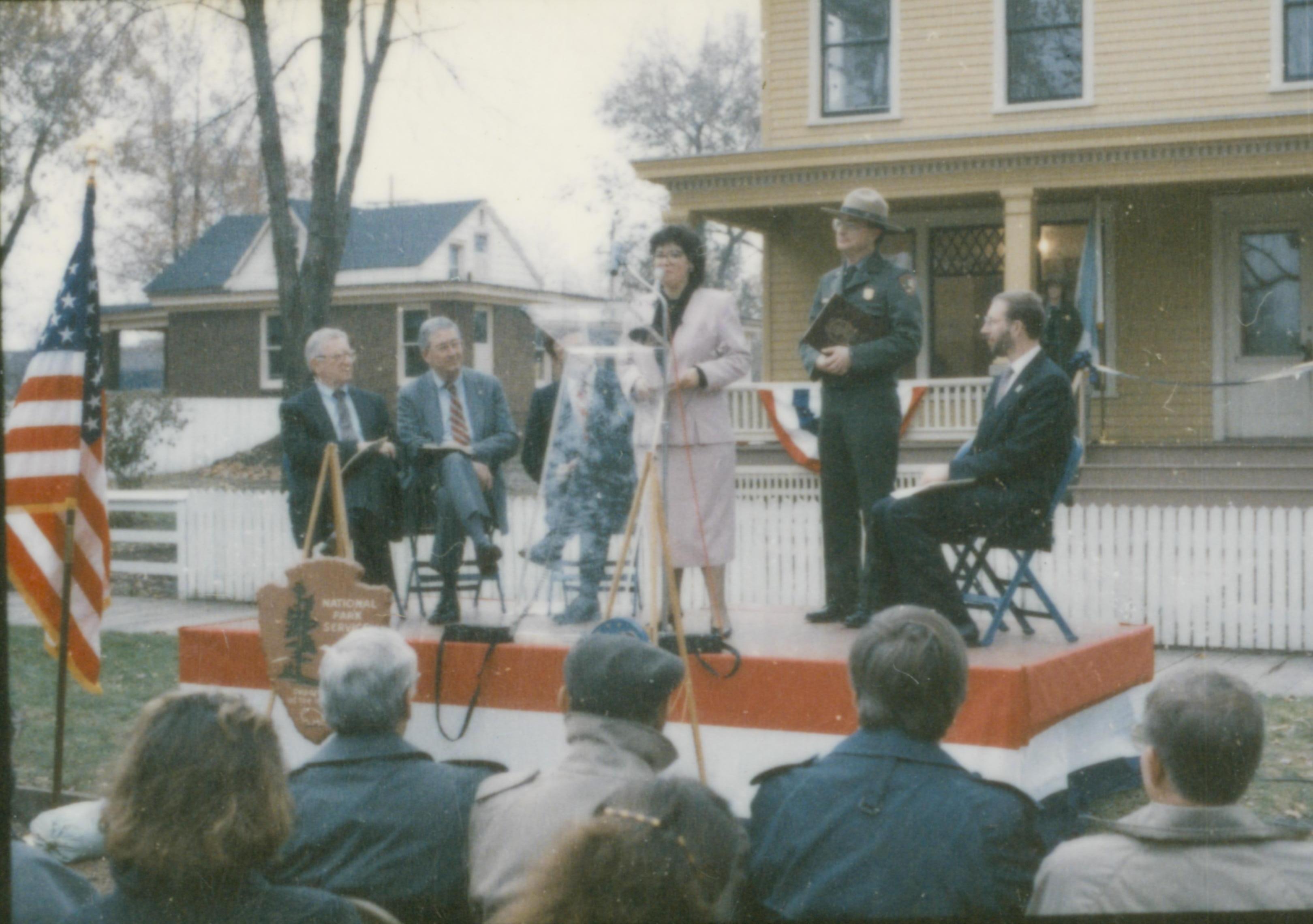 Five people on speakers platform in front of Cook house. Lincoln Home NHS- Cook House Re-opening, 1 Cook, ceremony