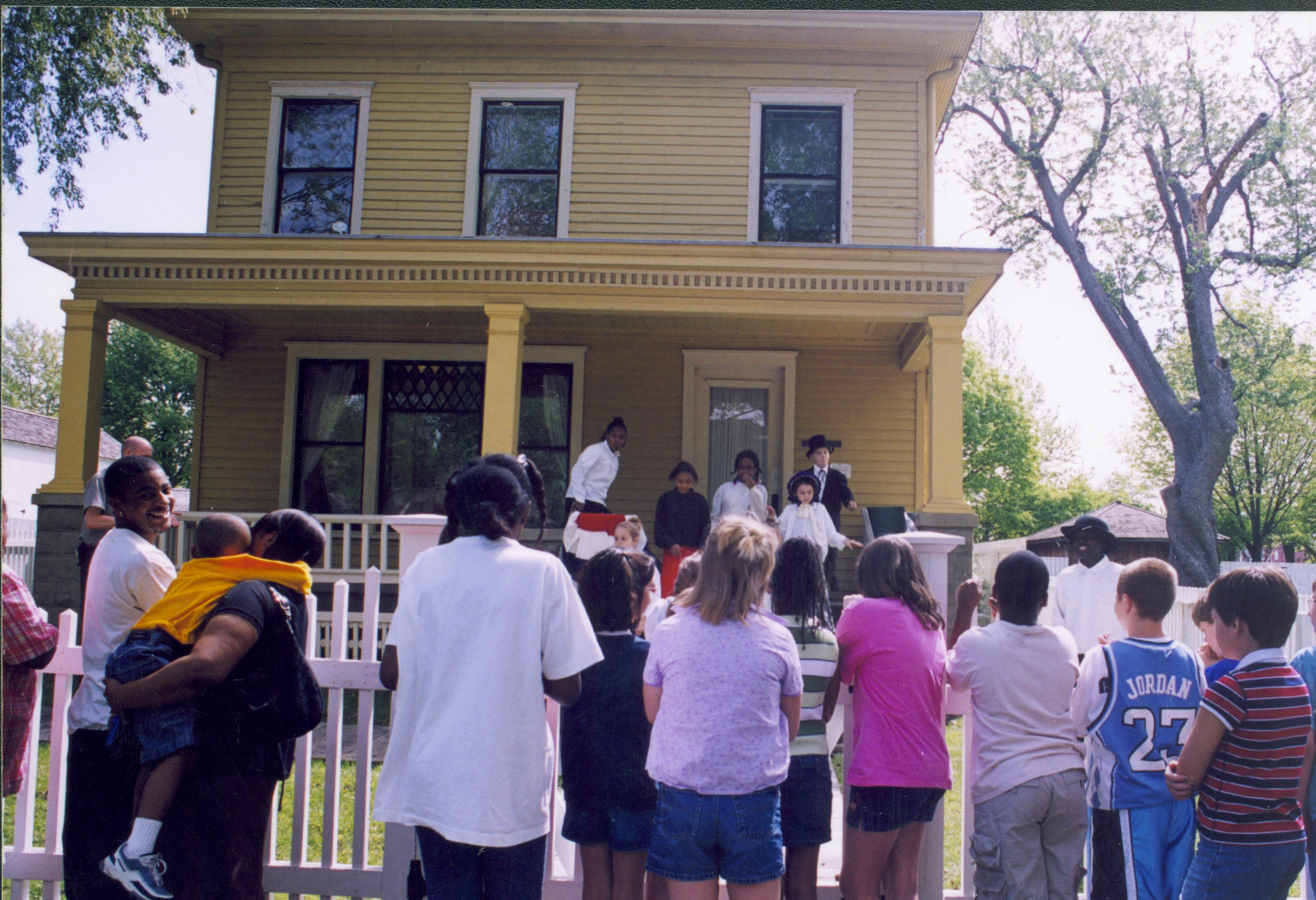 Dubois School Living History program - students perform skit on Women's Rights from Cook House porch while a school group watches.  Ranger Matt Brown just visible on left in front of porch. Arnold Barn visible on far left. Robinson Shed in background right. Looking East from 8th Street Dubois School, living history, Cook Robinson Shed, Arnold Barn, staff, students, school groups