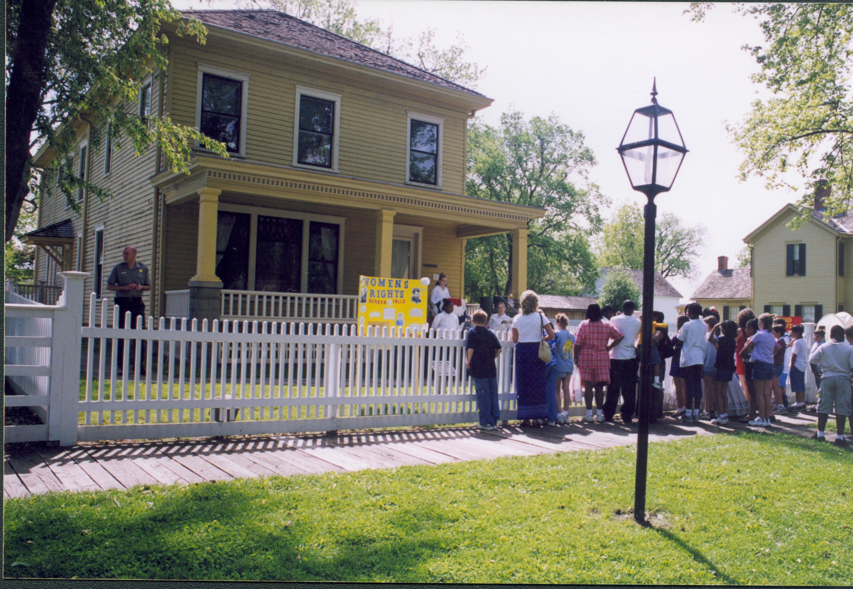 Dubois School Living History program - students perform a skit on Women's Rights sign on the Cook House porch while school groups and parents watch. Ranger Matt Brown observes on left. Allen Barn visible in background right, near Robinson House on far right.  Looking Southeast from 8th Street Dubois School, living history, staff, Cook, Robinson, Allen Barn, students, school groups