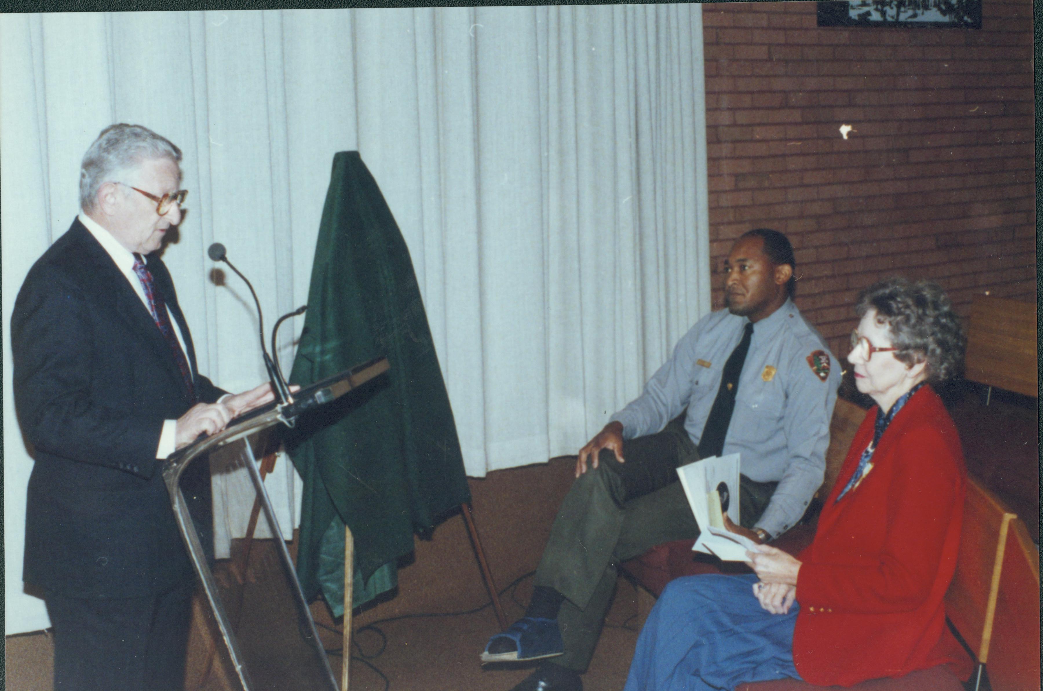 Man speaking into microphone, two people seated. Lincoln Home NHS- Bring Furniture Back Home ceremony, presentation