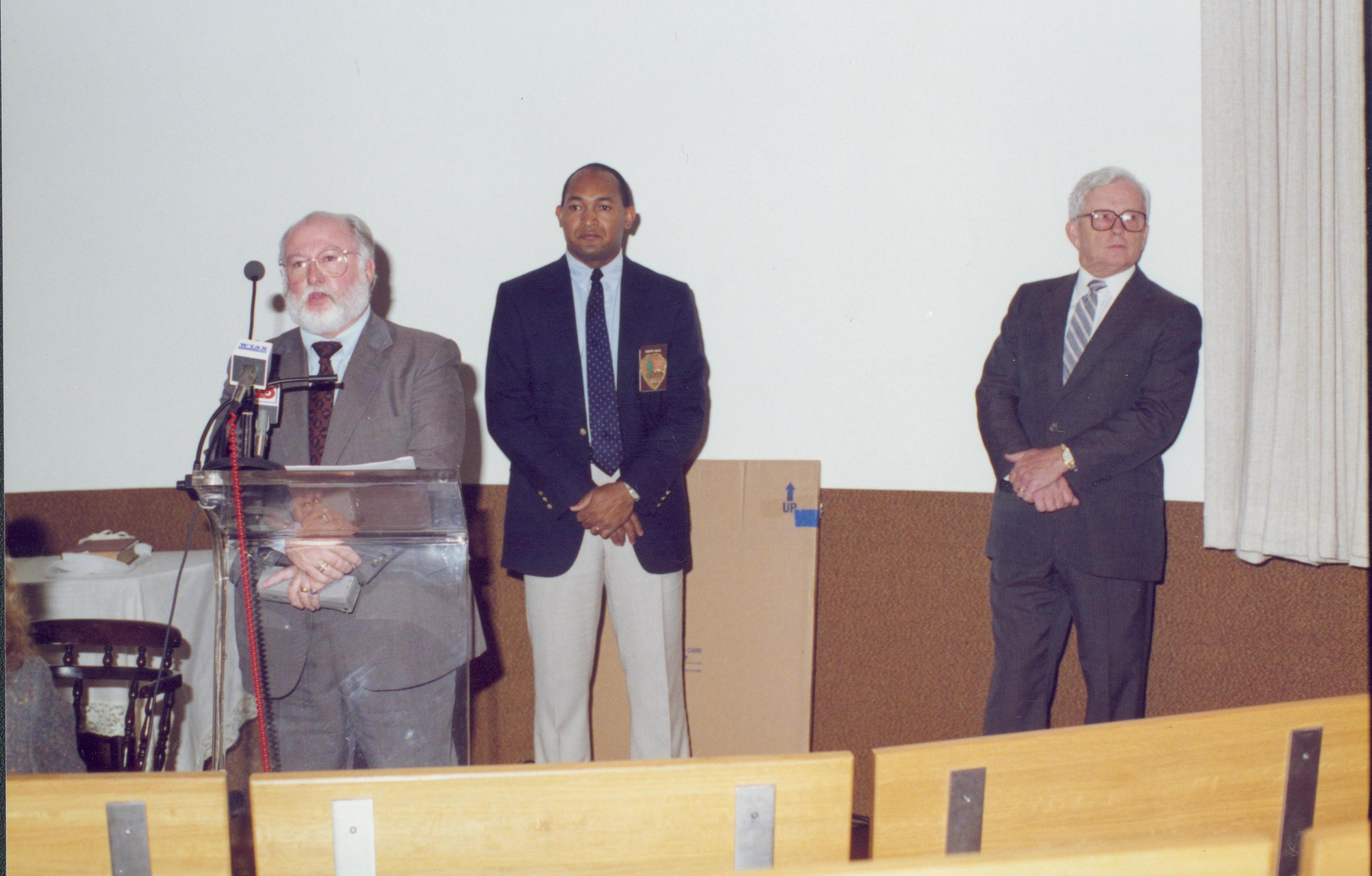 Man speaking into microphone, two people listening. Lincoln Home NHS- Bring Furniture Back Home ceremony, presentation