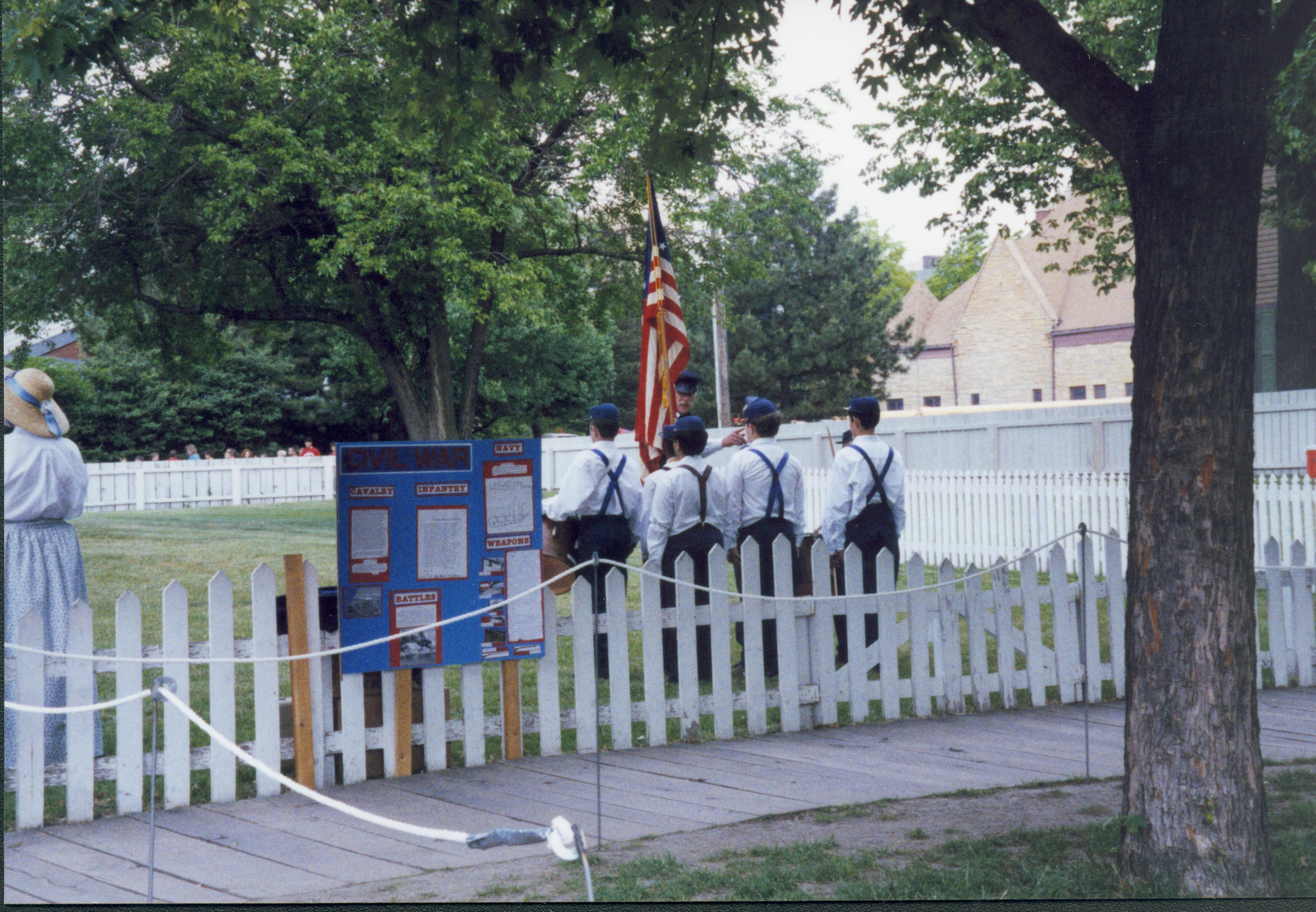 Iles School Living History Program - Students drilling in Civil War uniform in Burch Lot (Block 7, Lot 9). Mentor giving orders while holding US flag. Teacher/chaperone stands on left behind fence. School groups walk along alley in background left near Visitor Center. Grace Lutheran Church in background right.  Looking Northwest from 8th Street. Iles School, living history, Burch, Civil War, soldiers, Grace Lutheran Church, students, school groups 