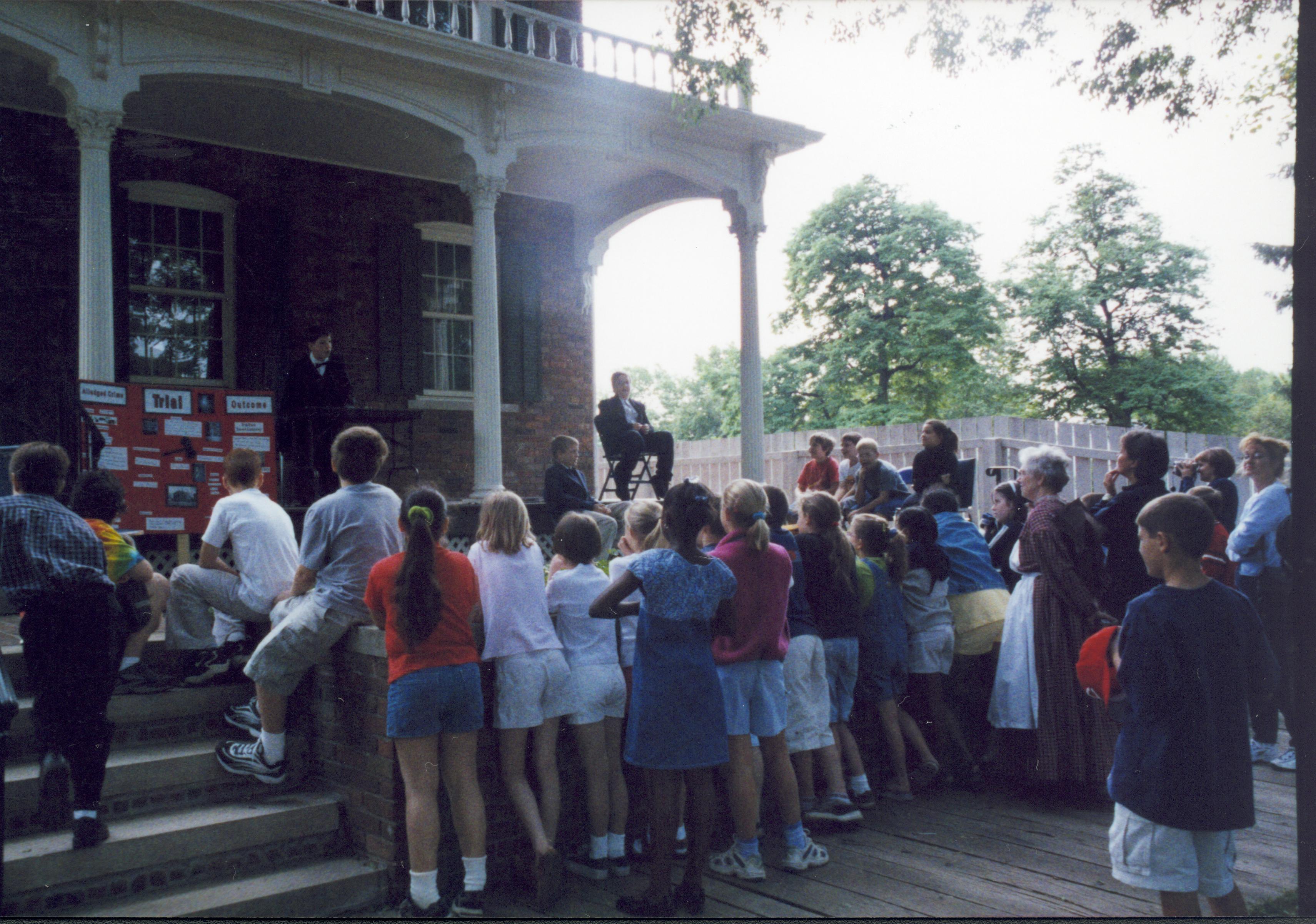 Iles School Living History program - Two boys on Conference Center porch perform a mock trial of one of Lincoln's cases. School groups watch from steps and boardwalk. Mentor in period clothing on right also watches performance. Looking Southeast from 8th Street Iles School, living history, Lincoln Legal Career, students, school groups, Conference Center
