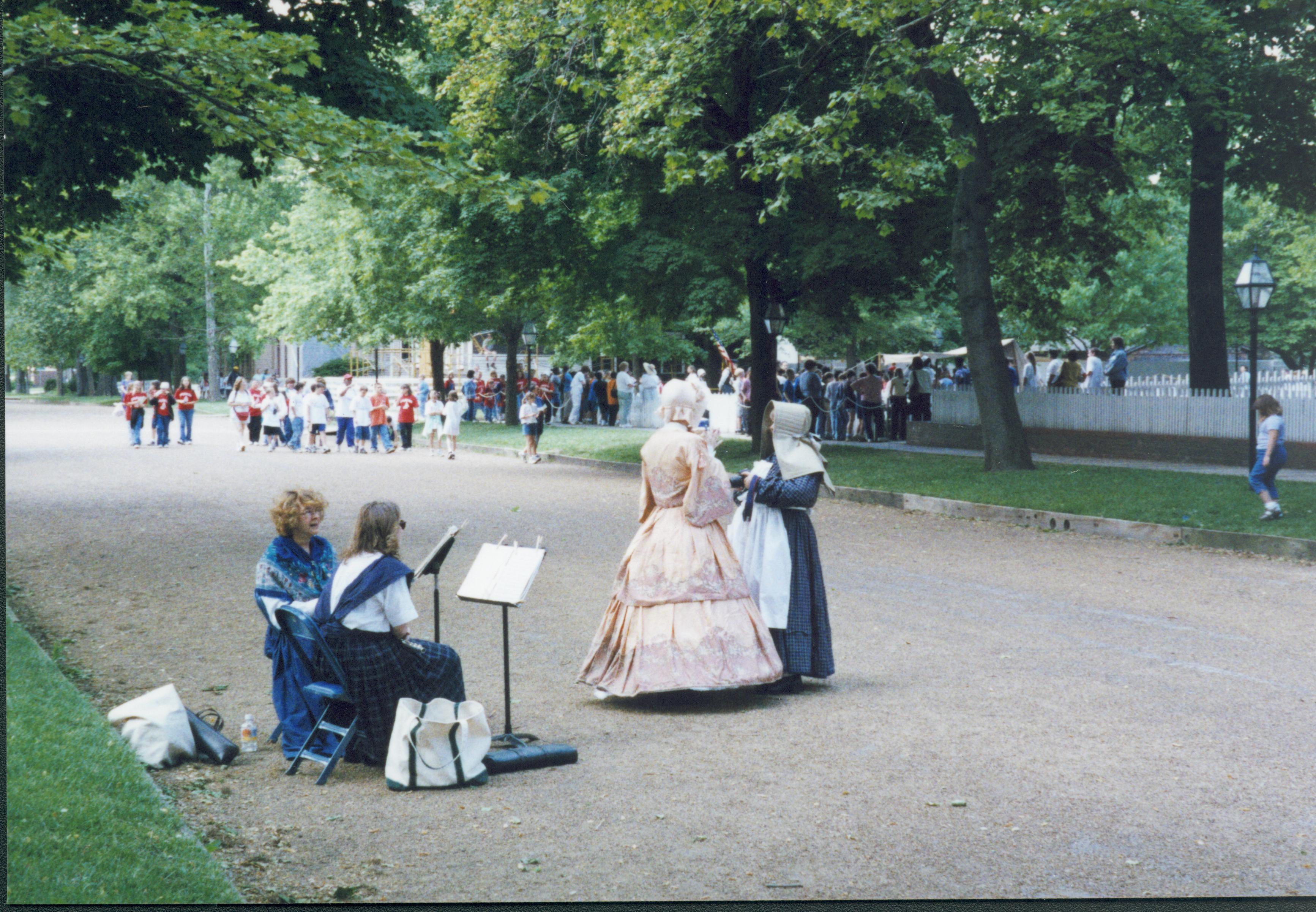Iles School Living History program - Two musicians sitting behind music stands in 8th Street while teachers in period dress discuss event.  School groups seen throughout park in background. Scaffolding for Corneau House in far background center. Looking Southwest from East side of 8th Street Iles School, living history, musicians, teachers, school groups, 8th Street, Corneau