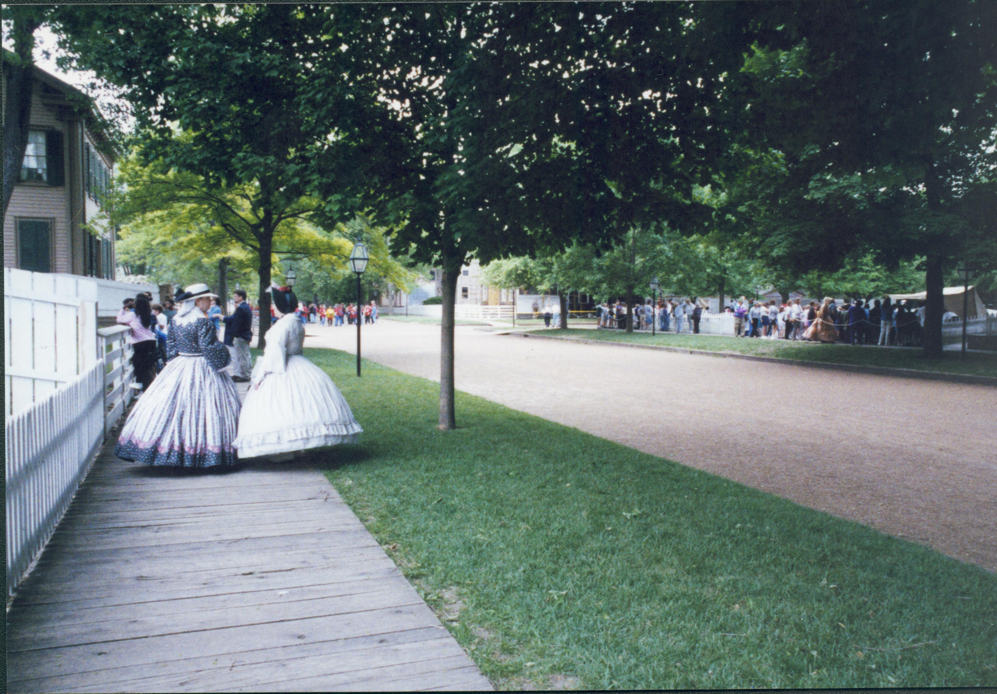 Iles School Living History program - Program Coordinators Paula Shotwell and Margie Atkins in period clothing chat on boardwalk. School groups throughout the park watching performances.  Lincoln Home in background left. Corneau and Miller houses just visible in far background center. Looking south from boardwalk on East side of 8th Street Iles School, living history, school groups, Lincoln Home, Corneau, Miller, 8th Street