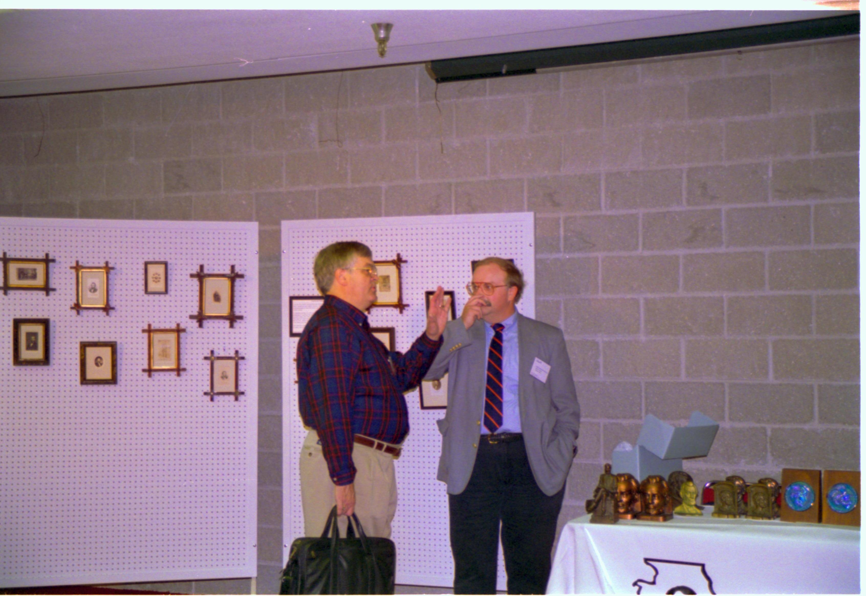 Two men talking in front of pictures hanging on peg board. 4-1997 Colloq (color); 5 Colloquium, 1997