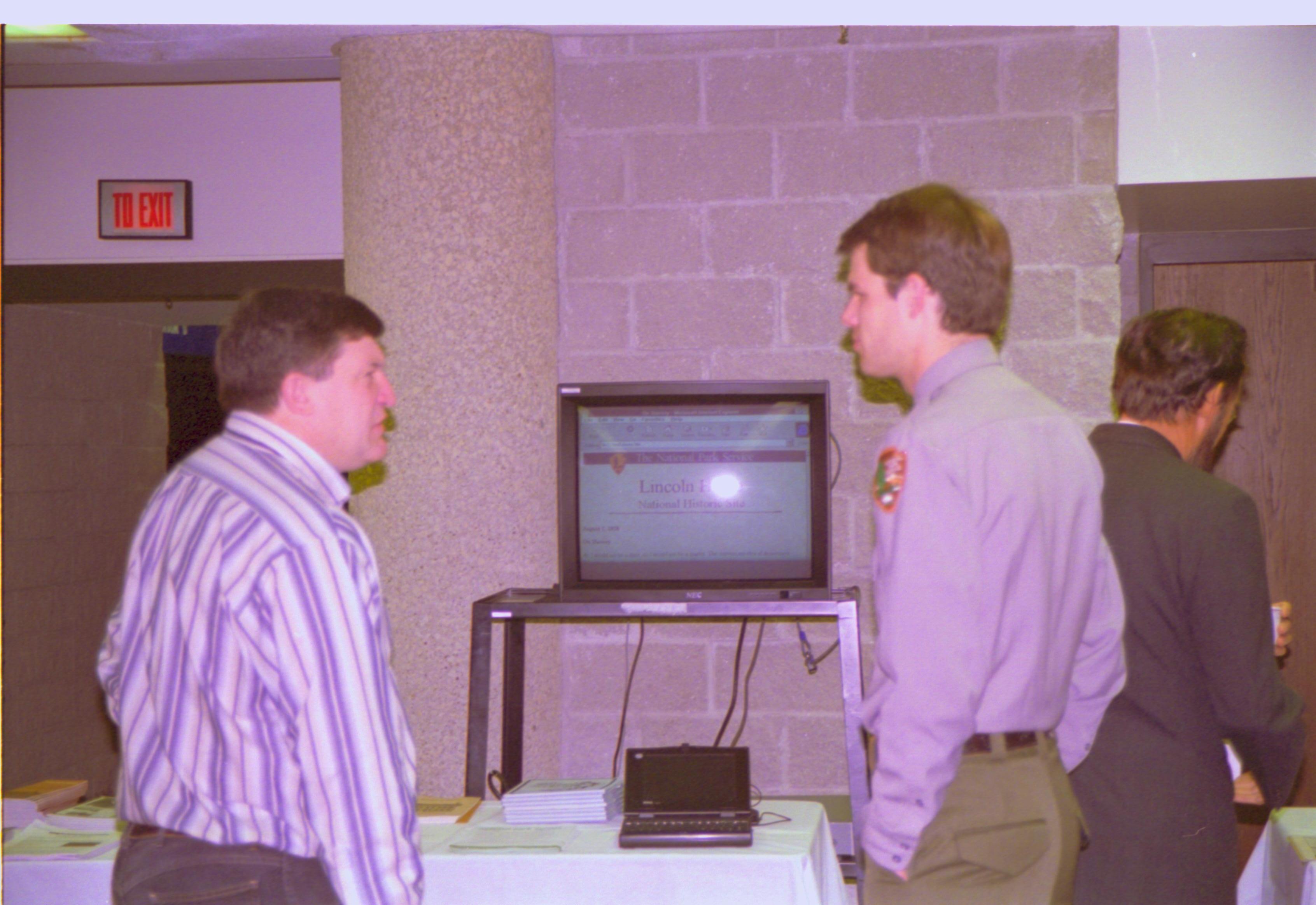 Two men talking in front of TV monitor. 4-1997 Colloq (color); 11 Colloquium, 1997