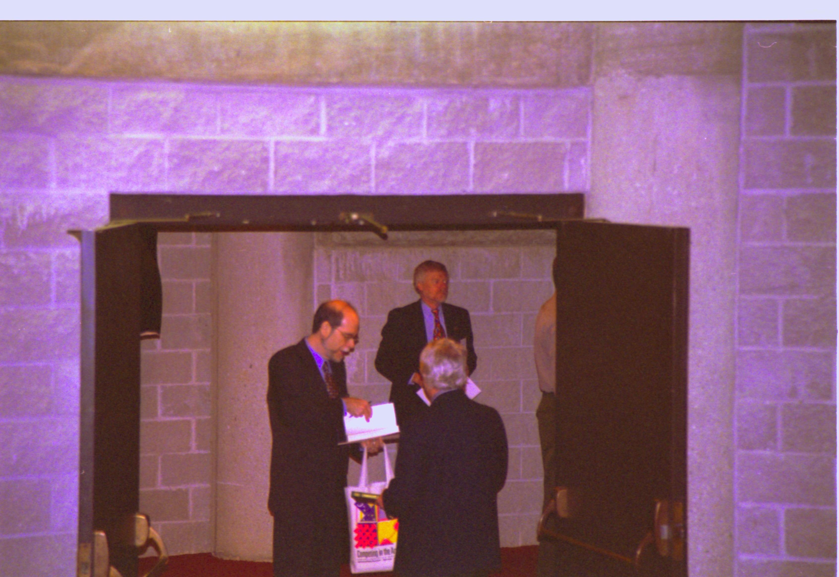 Three men standing in doorway, one looking at book. 3-1997 Colloq (color); 6 Colloquium, 1997