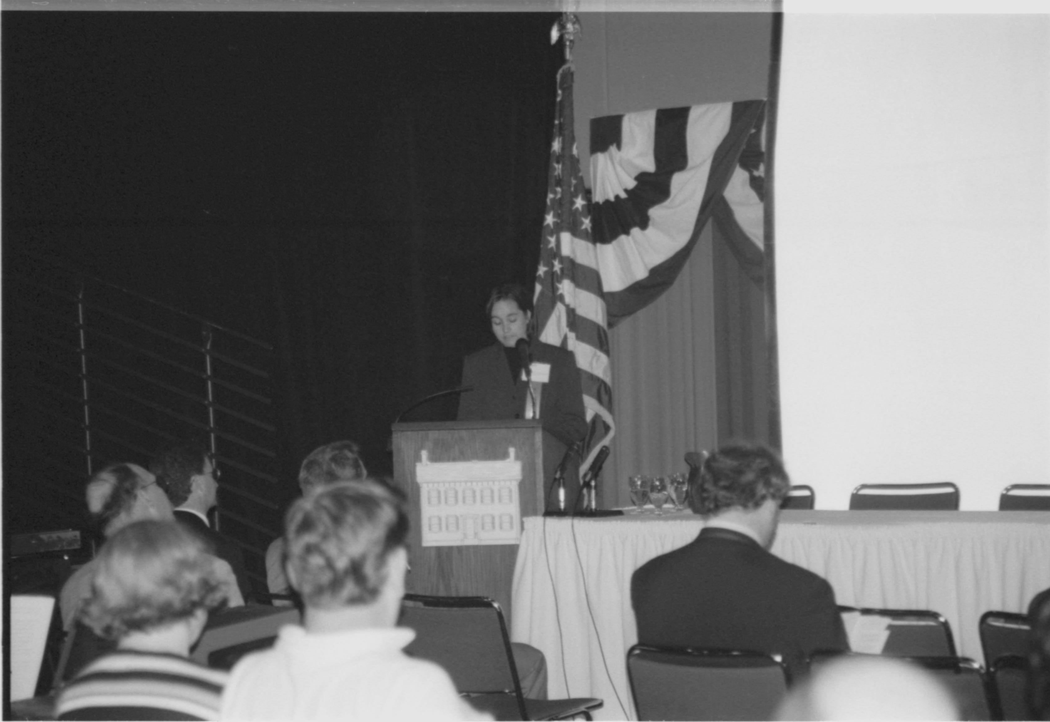 Lady speaking at podium, looking at notes. 1999-16; 27 Colloquium, 1999