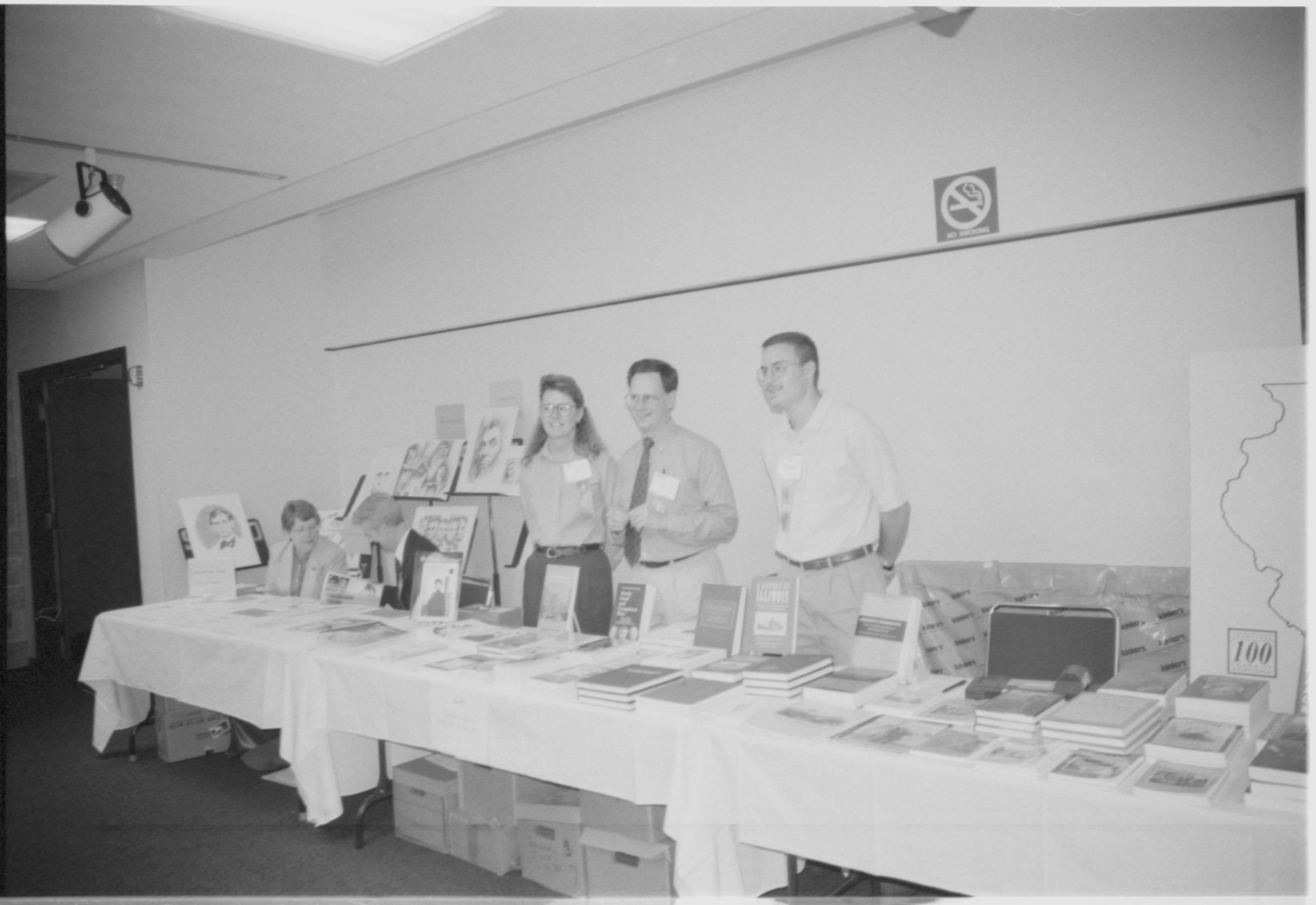 Three people standing behind exhibit table. 1999-16; 31 Colloquium, 1999