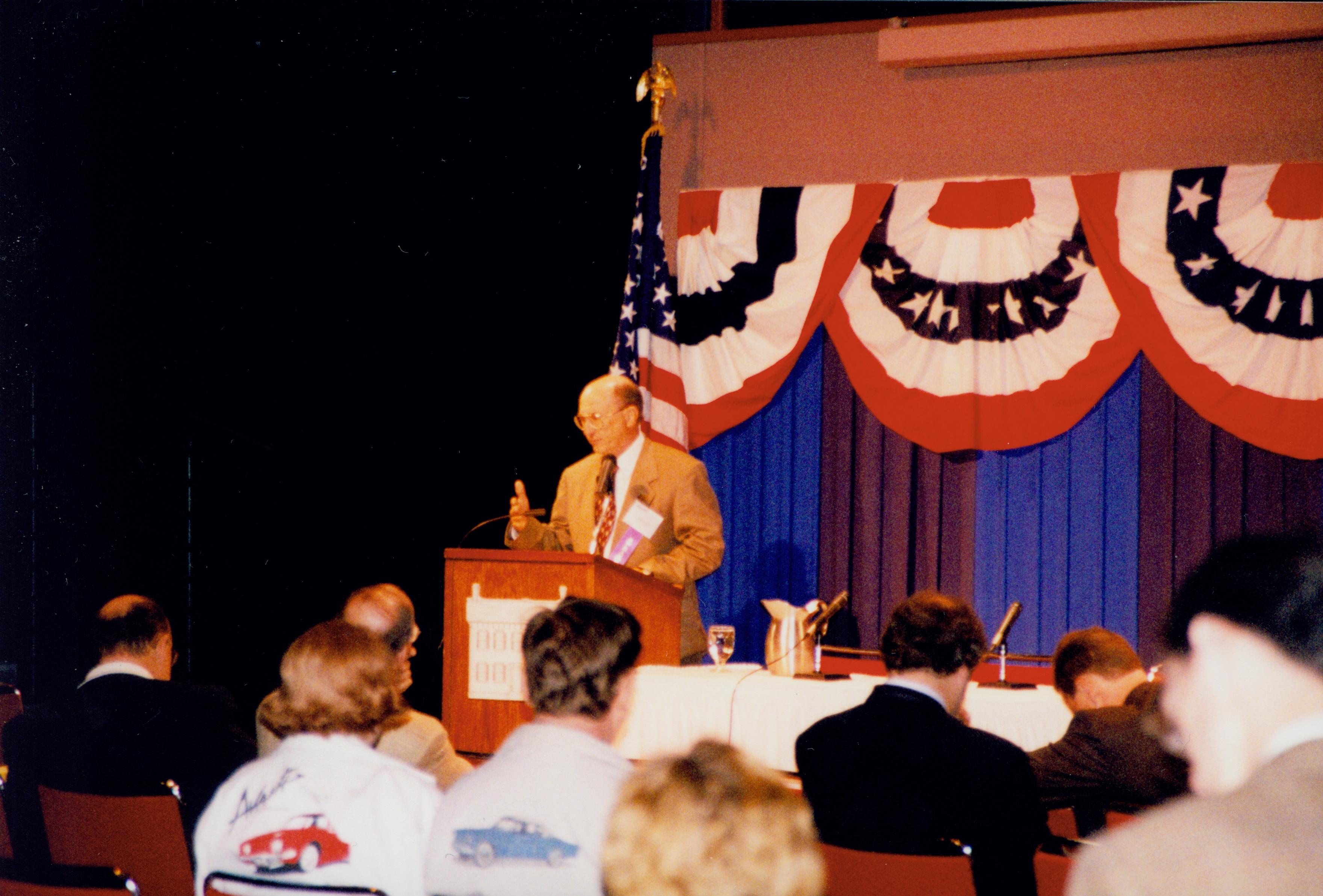 Man in tan coat speaking at podium. 1999-12; 4 Colloquium, 1999