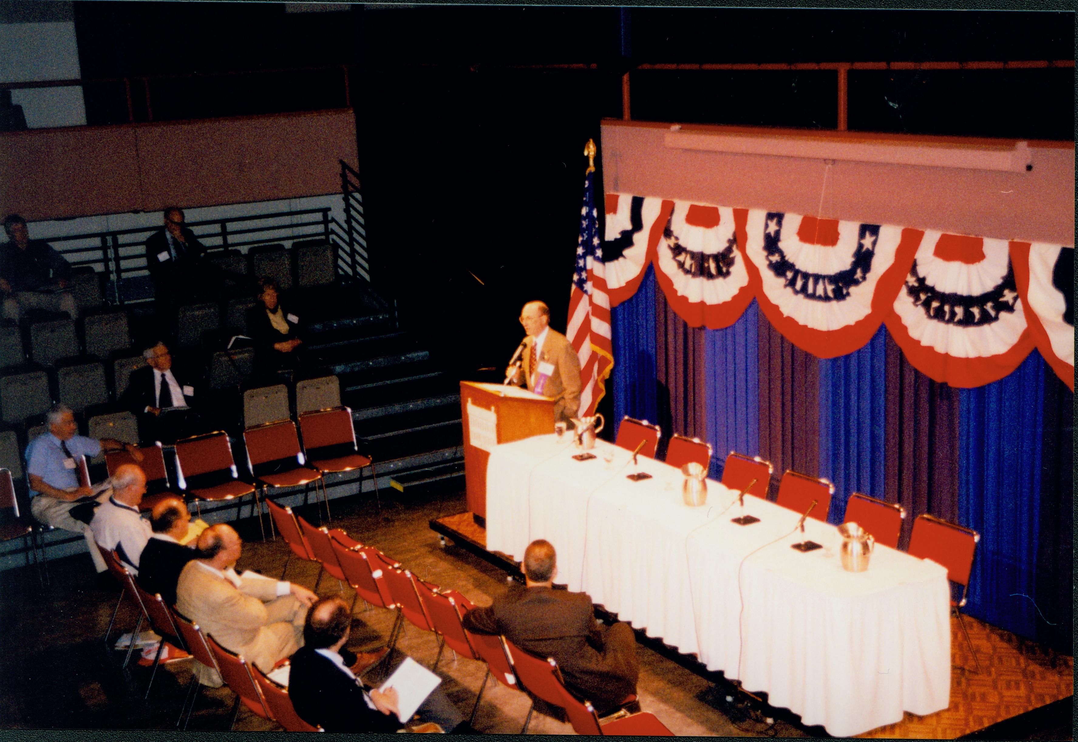 Man in tan coat speaking at podium (distant shot). 1999-12; 5 Colloquium, 1999
