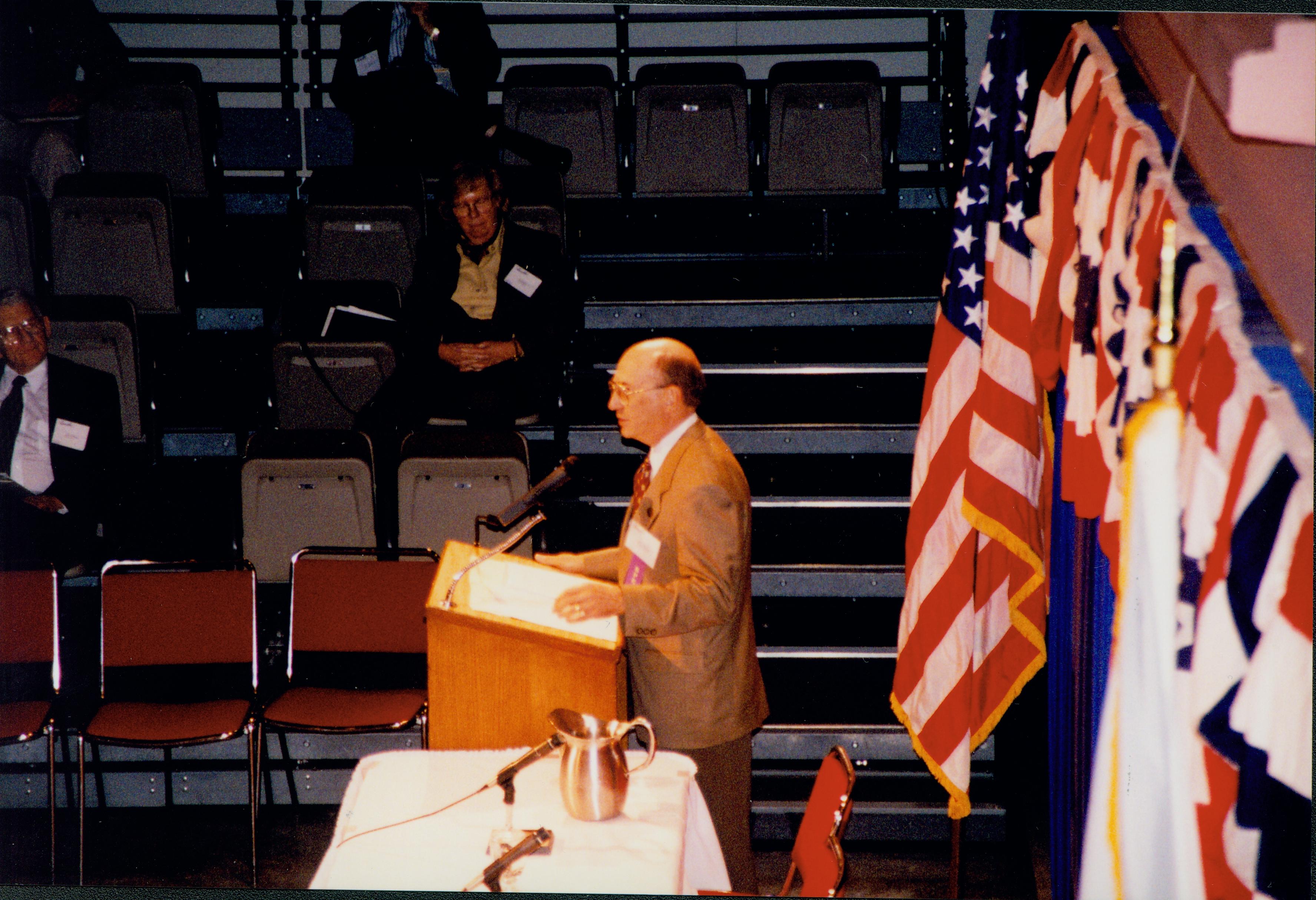 Man in tan coat speaking at podium (side view). 1999-12; 6 Colloquium, 1999