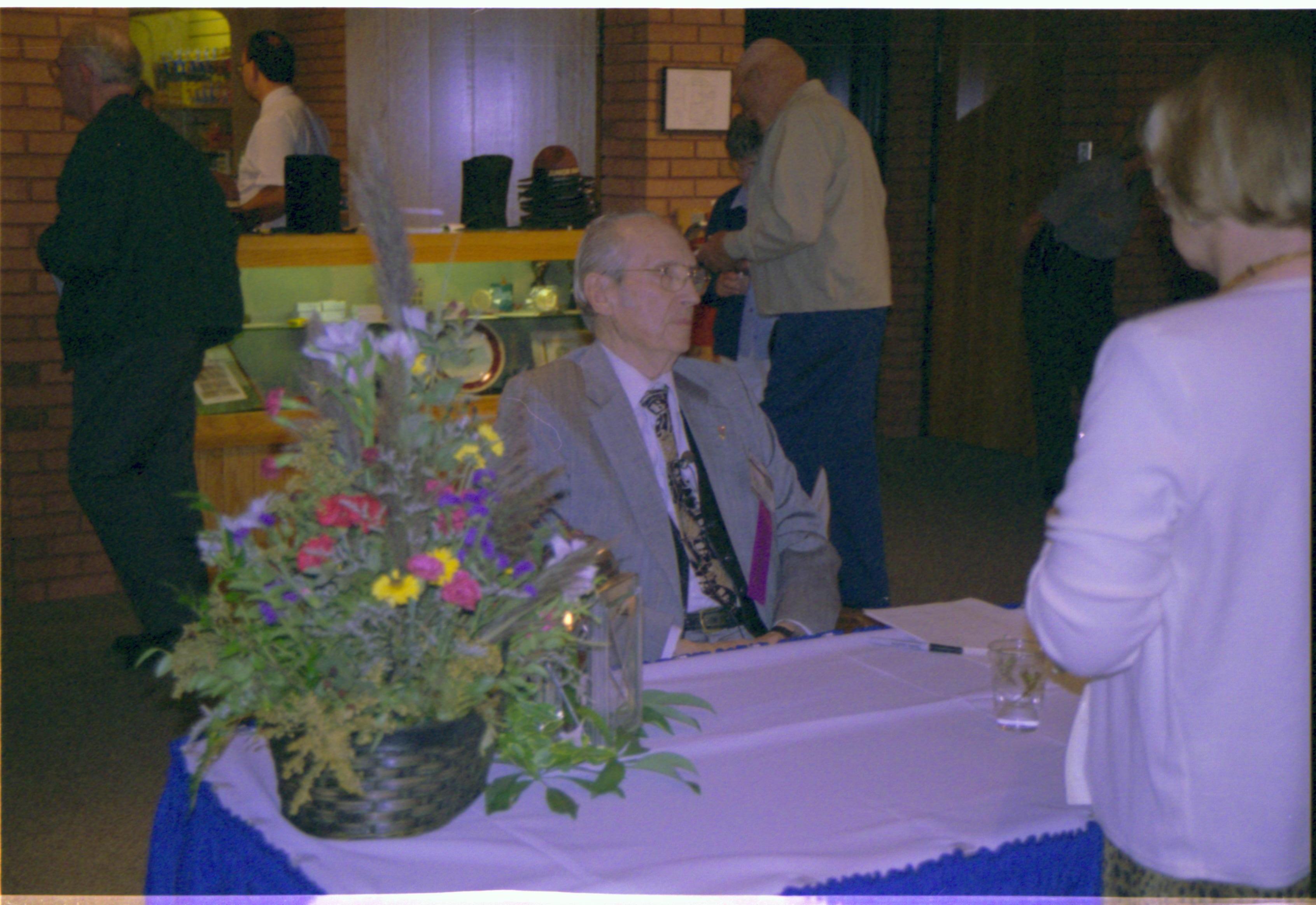 Man sitting at table with flower arrangement. Colloquium