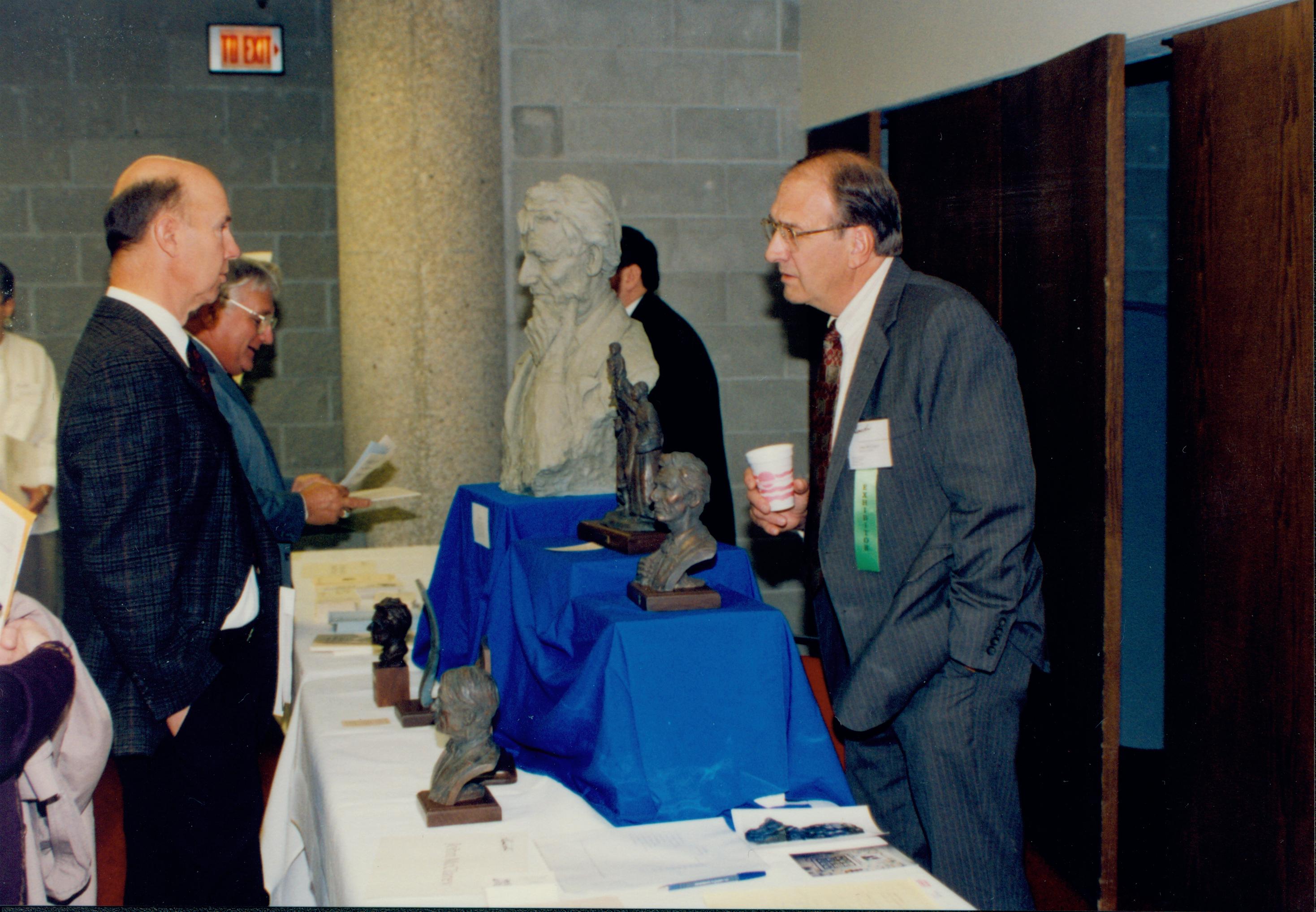 Man standing holding cup talking to another over sculptures.  Lincoln Colloquium; 56925 Lincoln, Colloquium