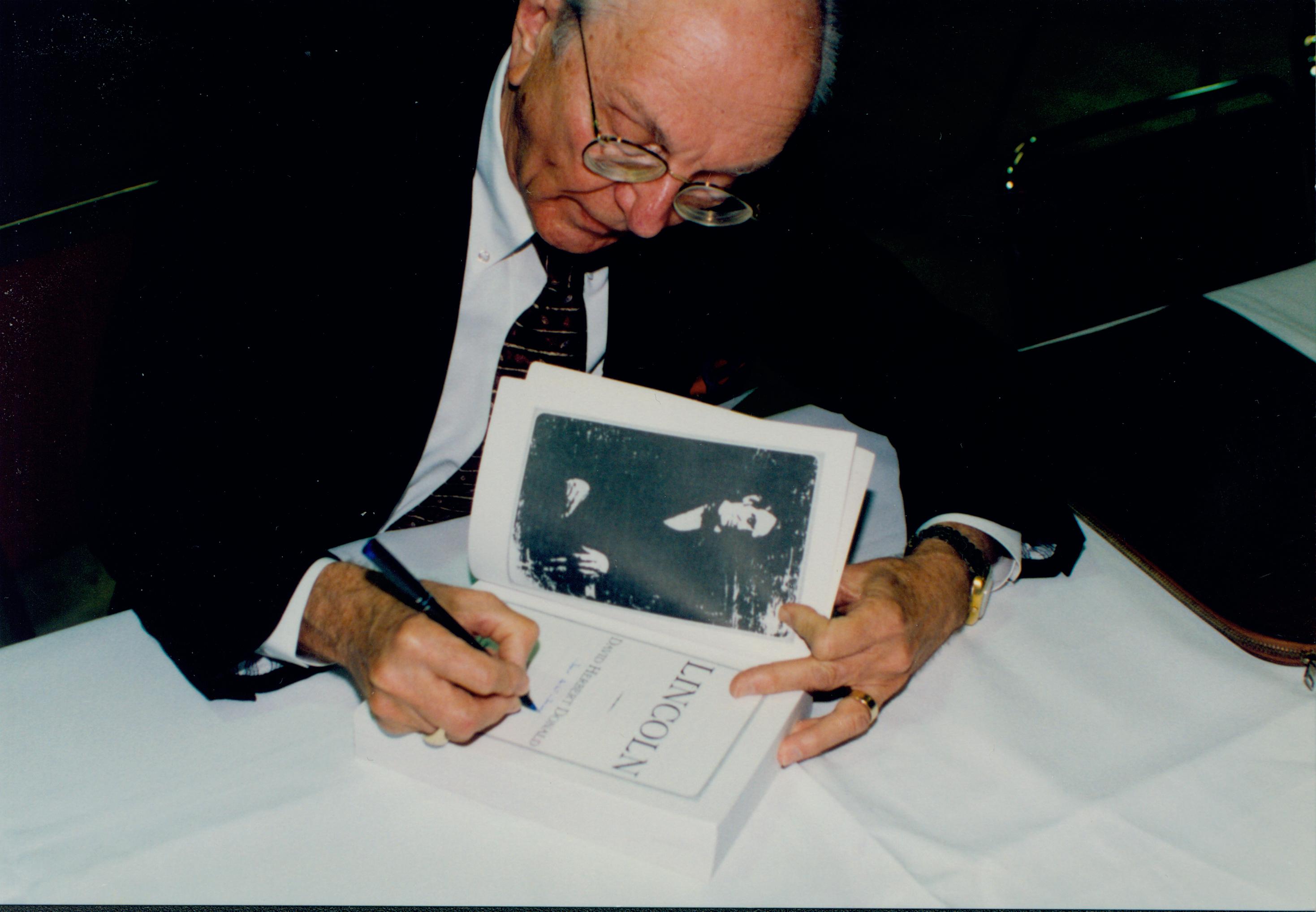 Man seated autographing book. Lincoln Colloquium; 56927 Lincoln, Colloquium