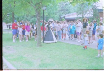 People standing around lady in period dress. Interpretation