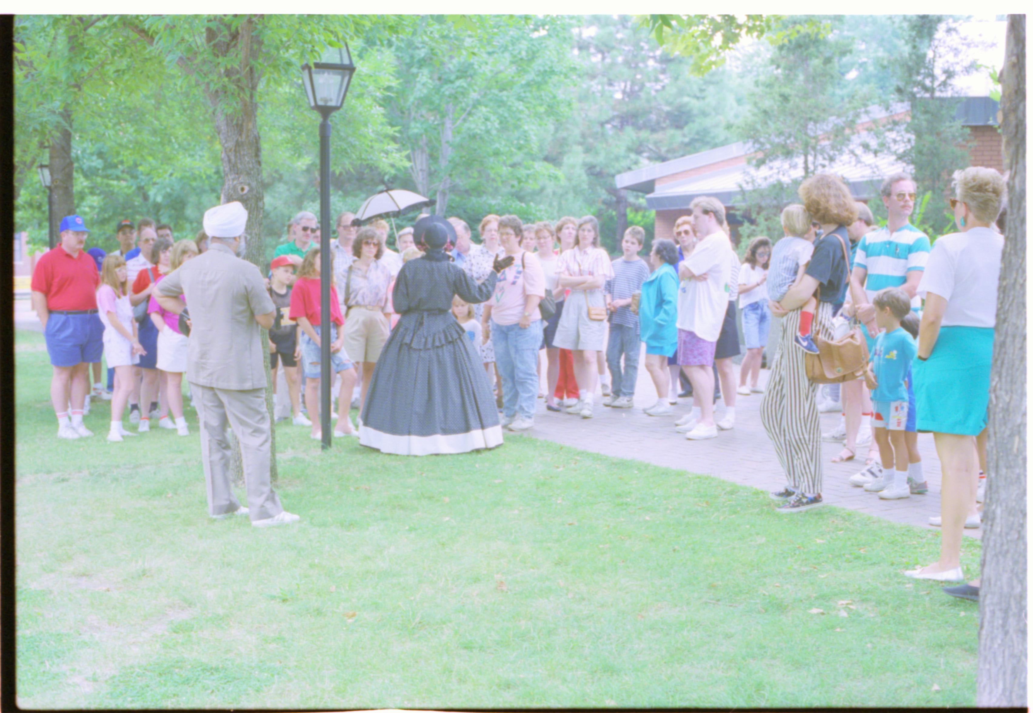 Group of people standing around lady in period dress. Interpretation