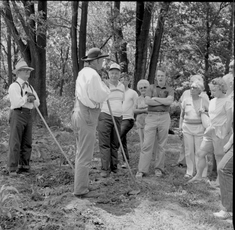 People in garden in period dress. Speaking to group. Log Cabin, State Park