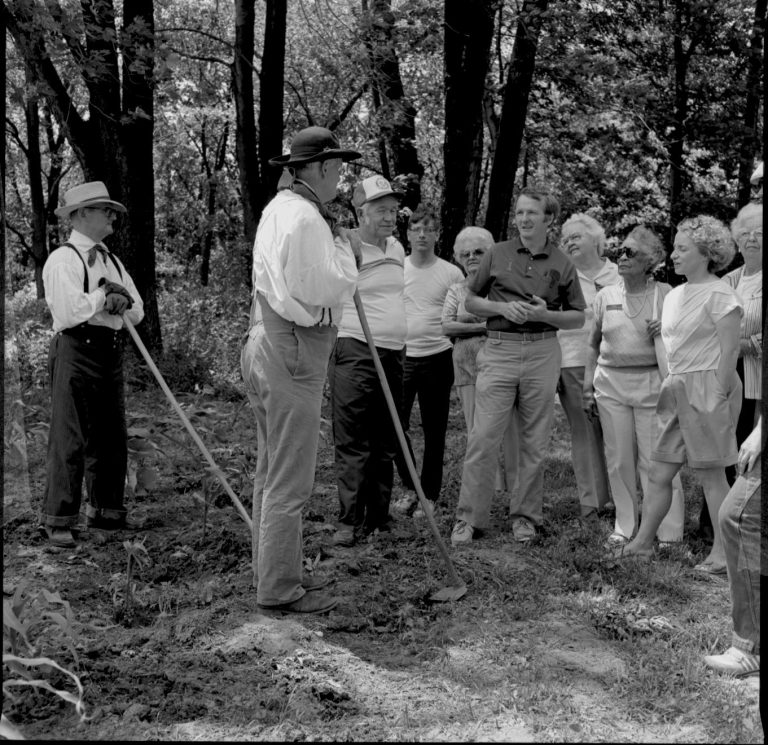 People in garden in period dress. Speaking to group. Log Cabin, State Park
