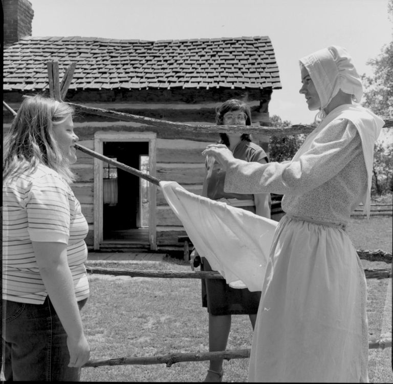 Lady in period dress hanging cloth. Log Cabin, State Park