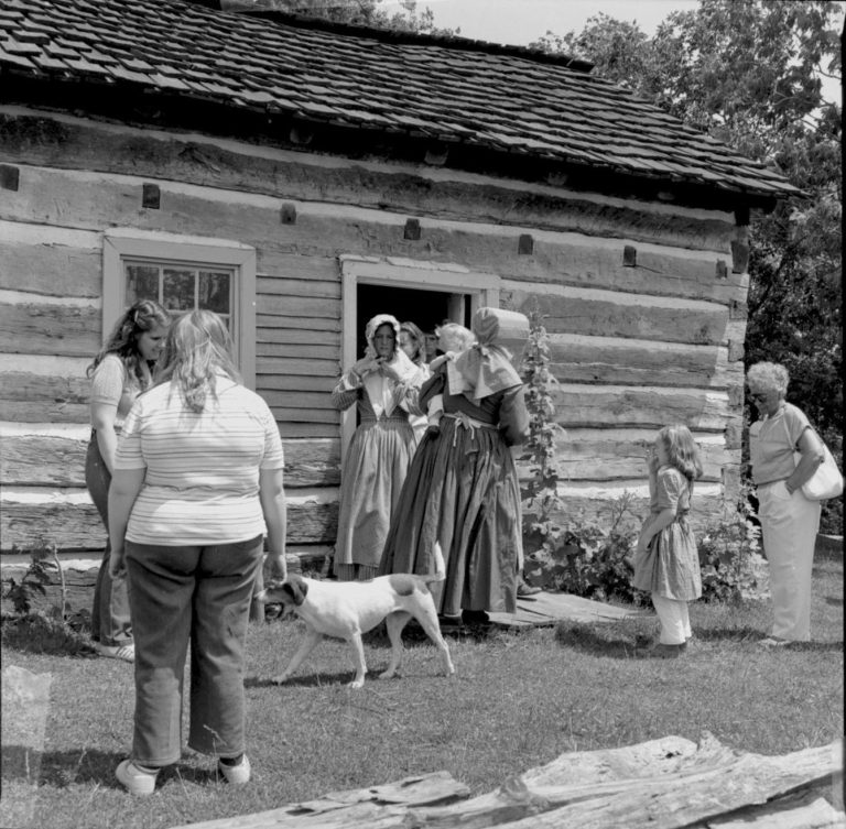 People in period dress outside log cabin.  Log Cabin, State Park