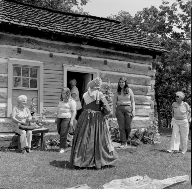 Lady in period dress outside log cabin. Log Cabin, State Park