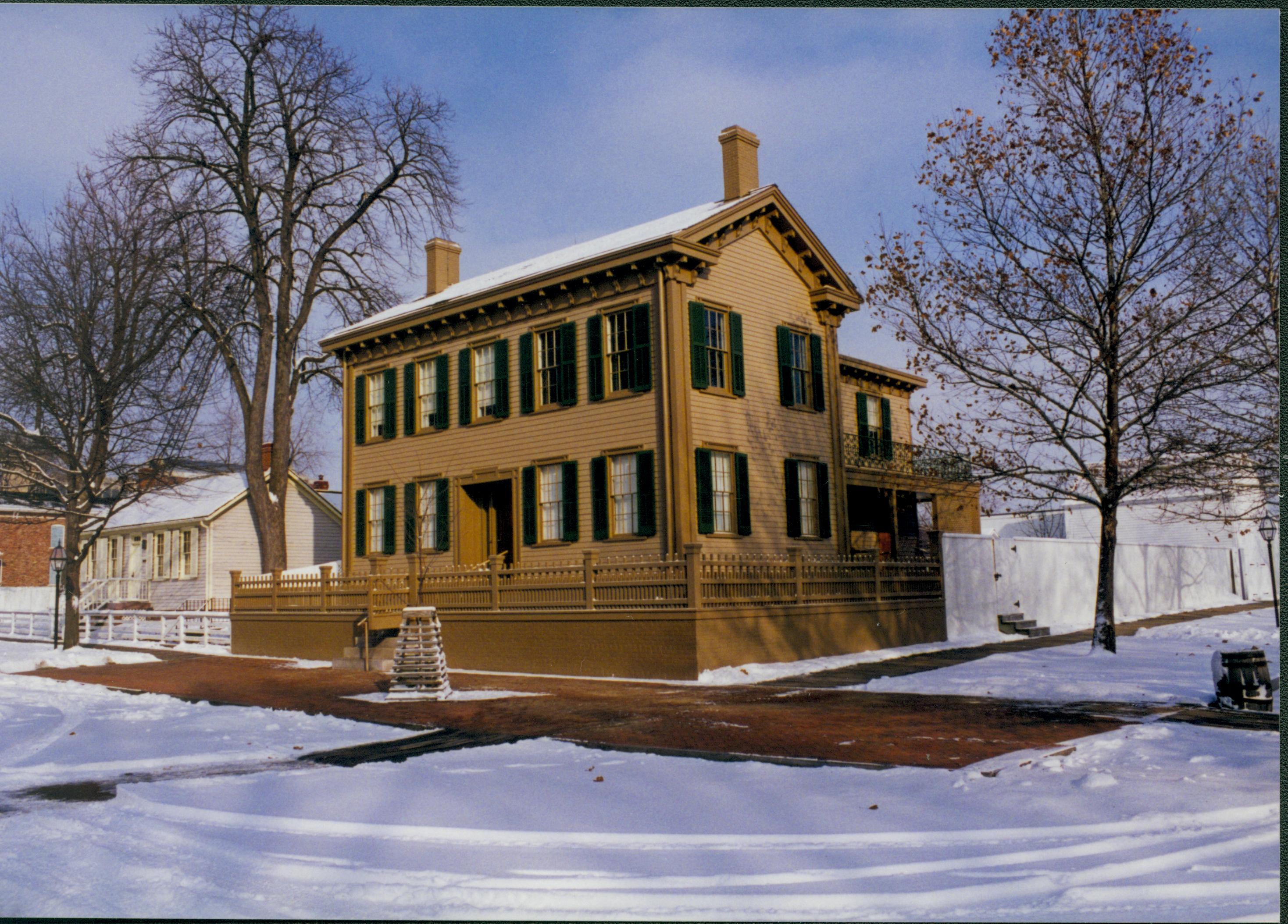 Lincoln neighborhood in the snow.  Lincoln Home in center with cleared brick plaza in front and elm tree in cage on plaza, Corneau House on left and Conference Center in brick on very far left. Trash barrel in gutter along Jackson Street on right.  Maintenance vehicle tracks on street Looking Northeast from 8th and Jackson Street intersection snow, Lincoln Home, Corneau, brick plaza, elm tree, 8th Street, Jackson Street