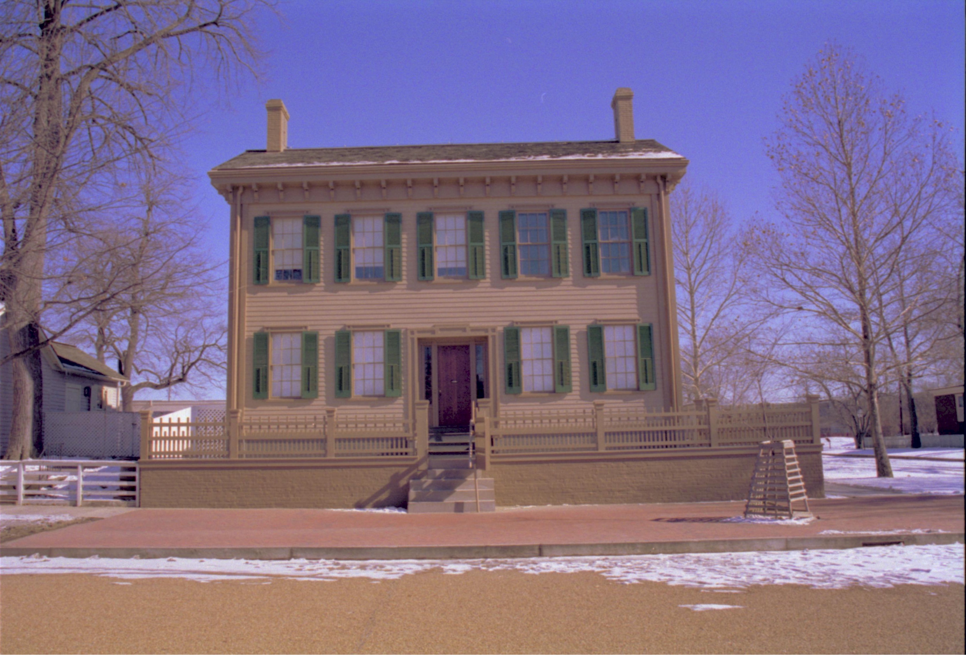 Lincoln Home in winter, some snow visible throughout. Cleared brick plaza in front with elm tree in cage on right.  Corneau House visible on left. Looking East from 8th Street snow, Lincoln Home, brick plaza, elm tree, Corneau, 8th Street