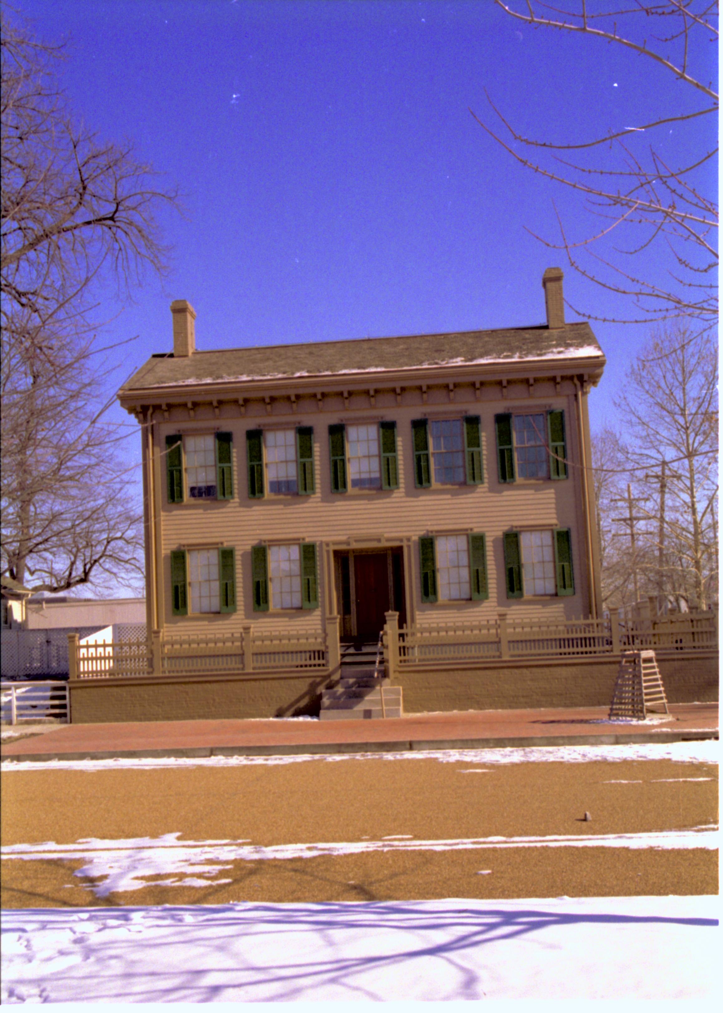 Lincoln Home in winter, some snow scattered throughout, cleared brick plaza in front, elm tree in cage on right.  B-2 in background left, Corneau House on far left. Looking East from boardwalk on west side of 8th Street snow, Lincoln Home, B-2, Corneau, brick plaza, elm tree, 8th Street