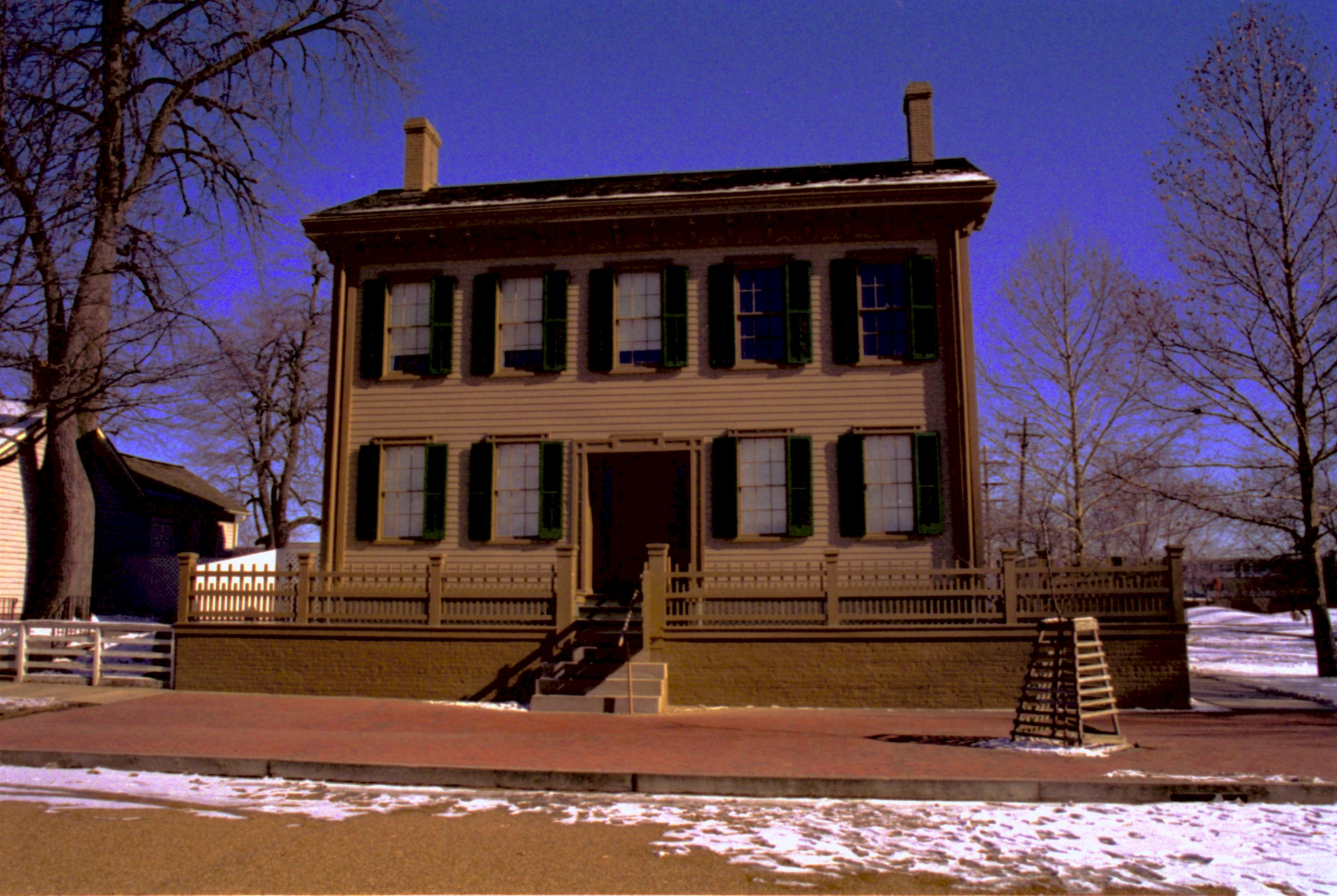 Lincoln Home in winter, some snow scattered throughout, cleared brick plaza in front, elm tree in cage on right.  Corneau House on left, former Travel Lodge Motel in far background right Looking East from 8th Street snow, Lincoln Home, brick plaza, elm tree, 8th Street, Corneau, Travel Lodge Motel