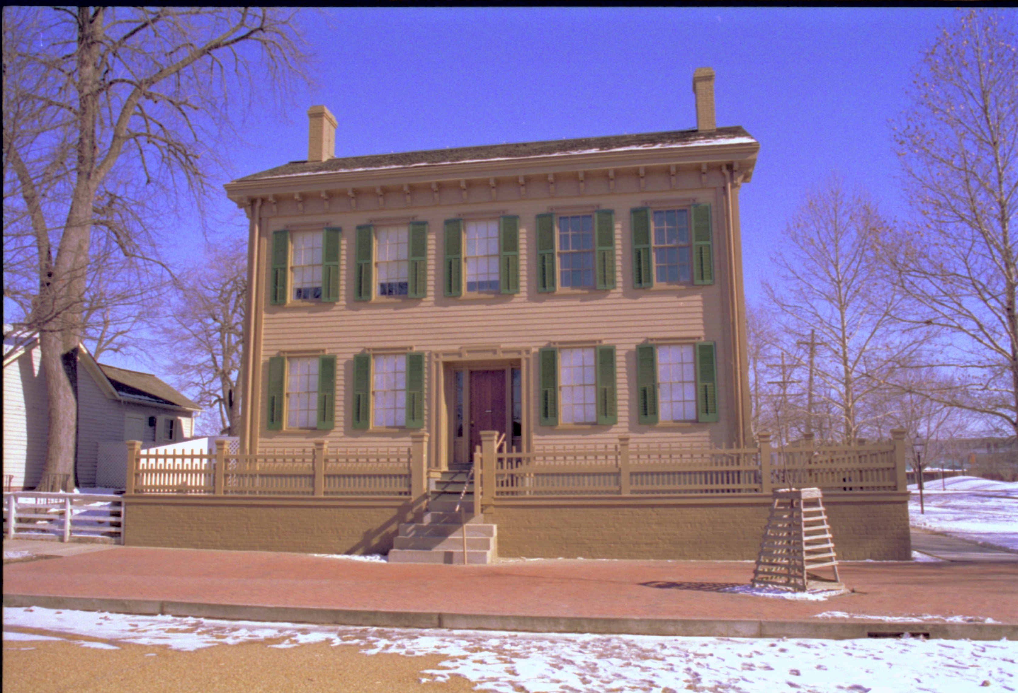 Lincoln Home in winter, some snow scattered throughout, cleared brick plaza in front, elm tree in cage on right.  Corneau House on left, former Travel Lodge Motel in far background right Looking Northeast from 8th Street snow, Lincoln Home, brick plaza, elm tree, Corneau, Travel Lodge Motel, 8th Street