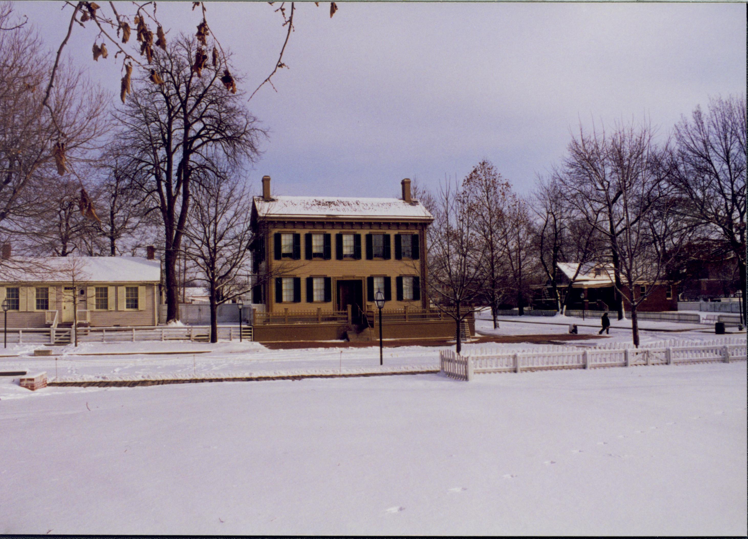 Lincoln neighborhood in the snow from west end of Brown lot.  Lincoln Home in center, Corneau house on left, Arnold House in background on right. Footprints and a few animal tracks visitble in snow. Visitor crossing Jackson Street near trash barrels   Looking east from Brown lot Block 7, Lot 10 snow, Lincoln Home, Corneau, Arnold, 8th Street, Jackson Street, visitor