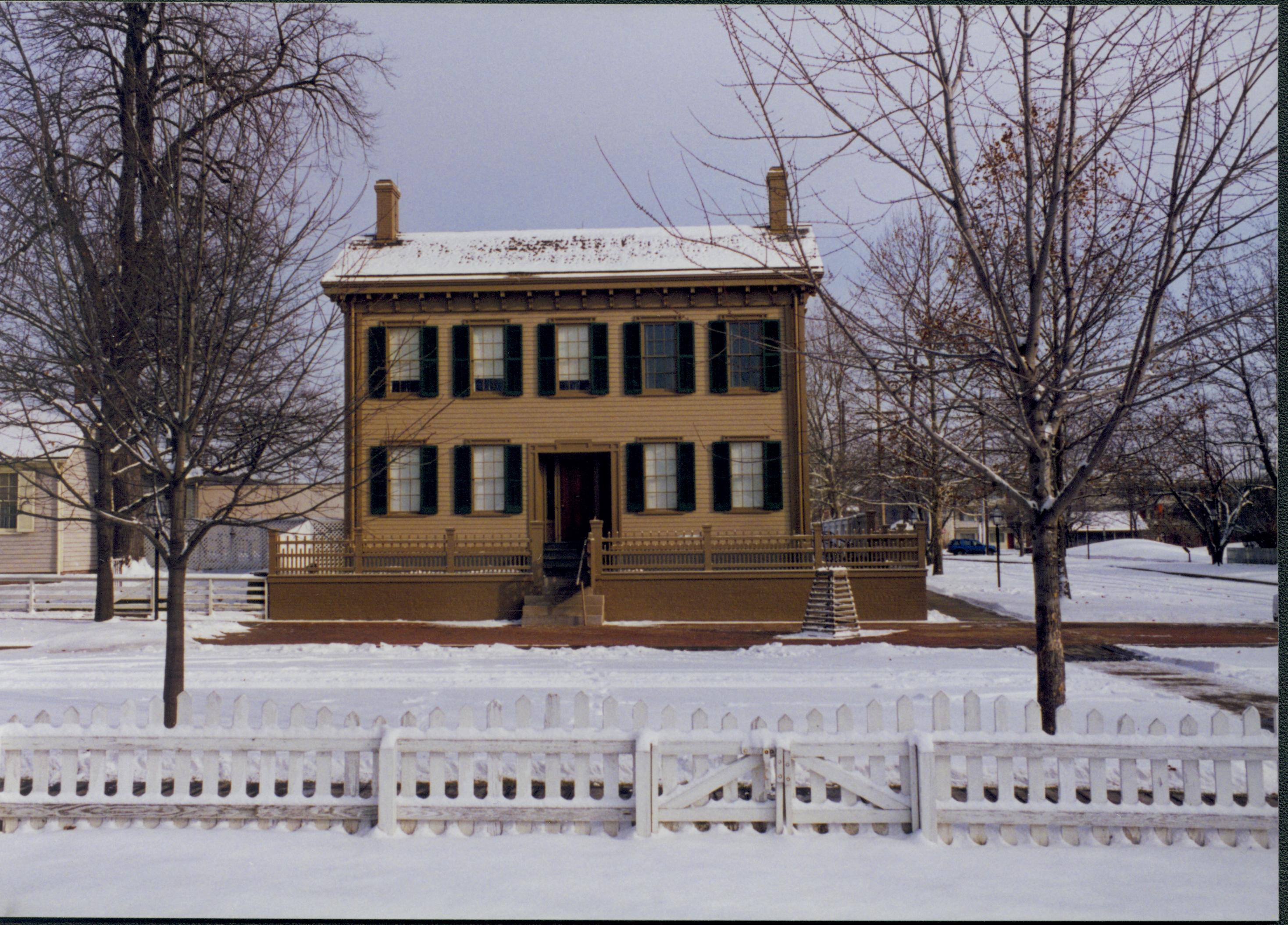 Lincoln Home in the snow.  Corneau House on the left, brick plaza with elm tree in front cleared of snow along 8th Street.  Fence in foreground surrounds Burch lot. Looking East from Burch lot, Block 7, Lot 9. snow, Lincoln Home, Corneau, 8th Street, brick plaza, elm tree