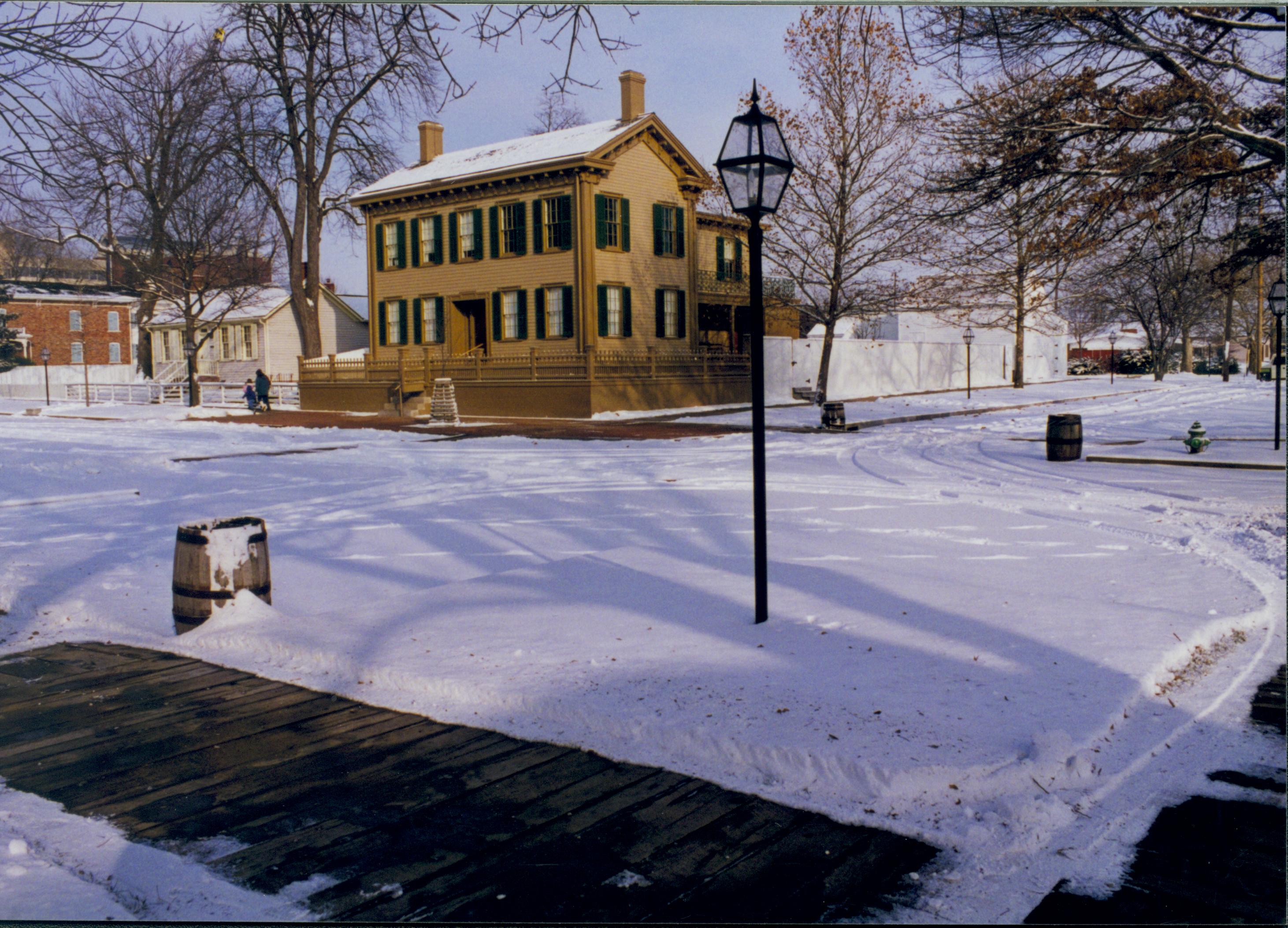 Lincoln neighborhood in the snow.  Lincoln Home in center with cleared brick plaza and elm tree in cage on plaza.   Corneau house on left, Conference Center in brick on far left.  Boardwalks cleared of snow in foreground with trash barrels visible on nearly every corner of intersection. Maintenance vehicle tracks and footprints visible in snow.  Visitors walk away from Lincoln Home. Looking Northeast from just off 8th and Jackson Street intersection  snow, Lincoln Home, Corneau, 8th Street, Jackson Street, brick plaza, elm tree, boardwalks, Conference Center