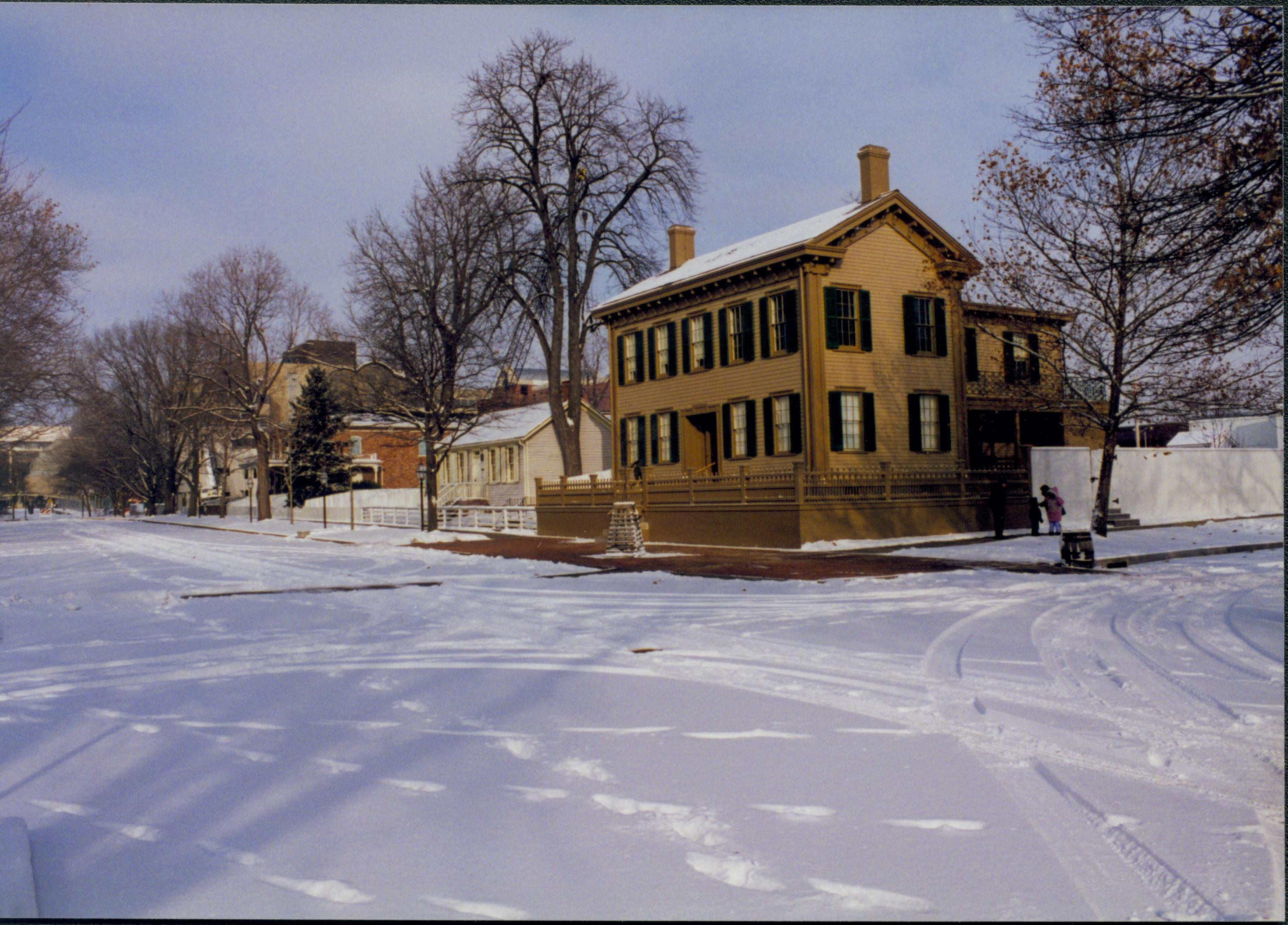 Lincoln neighborhood in the snow.  Lincoln Home in center with cleared brick plaza in front and elm tree in cage on plaza.  Corneau House on left and Conference Center on far left. Sangamon County Courthouse visible over Corneau roof.  Maintenance vehicle tracks and footprints visible in snow in 8th and Jackson Street intersection.  Visitors on boardwalk along Jackson Street near trash barrel. Looking Northeast from just past intersection of 8th and Jackson Streets snow, Lincoln Home, Corneau, Conference Center, Sangamon County Courthouse, 8th Street, Jackson Street, brick plaza, elm tree, visitors