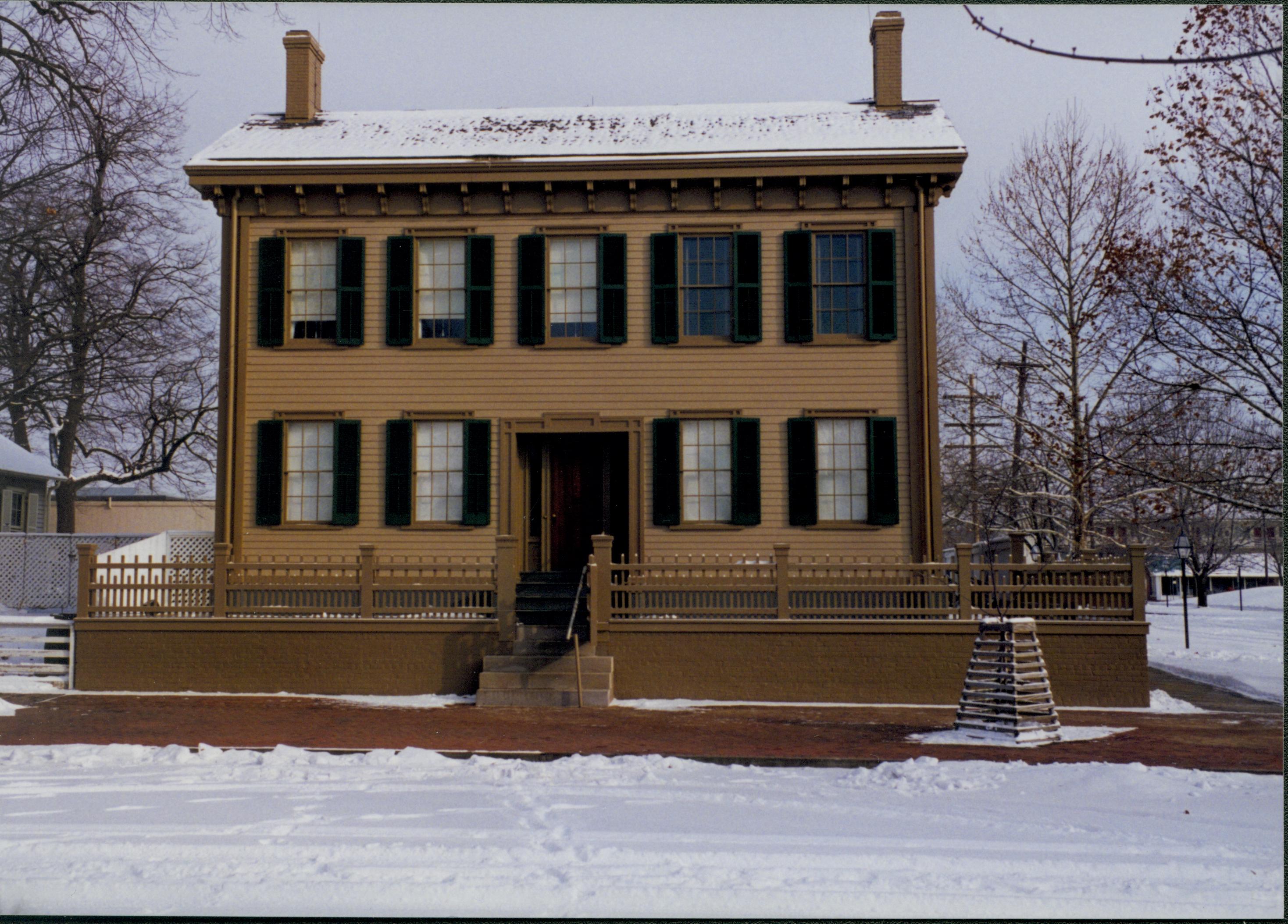 Lincoln Home in the snow with cleared brick plaza in front, elm tree in cage on right. Corner of Corneau House visible on far left.  Footprints and maintenance vehicle tracks visible in snow on 8th Street  Looking East from 8th Street snow, Lincoln Home, 8th Street, brick plaza, elm tree, Corneau