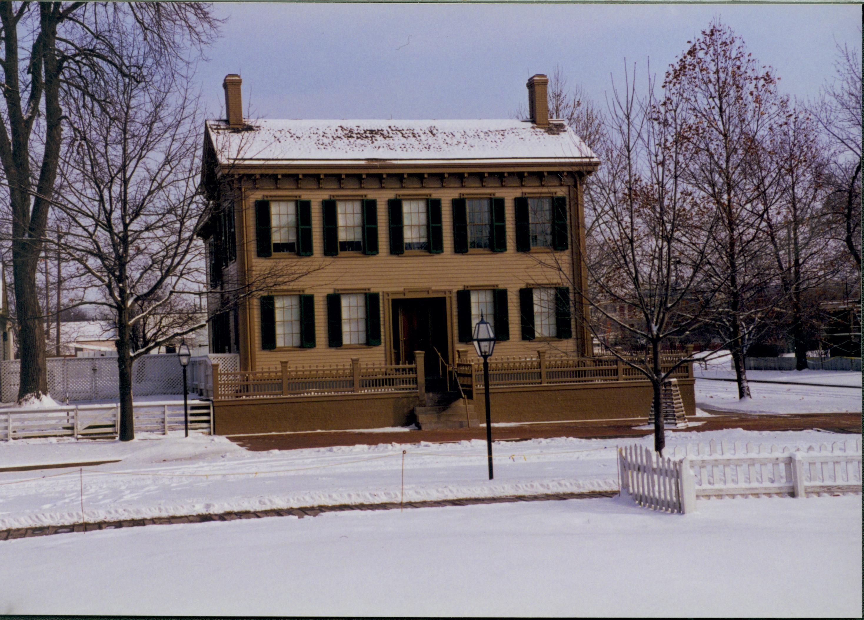 Lincoln Home in the snow, cleared brick plaza in front. Elm tree in cage on right, fences for the Carrigan lot on left, and Burch lot in foreground right. Back corner of Arnold House just visible on far right. Looking East from Brown lot, Block 7, Lot 10 snow, Lincoln Home, brick plaza, elm tree, fences, 8th Street, Carrigan, Brown lot, Arnold