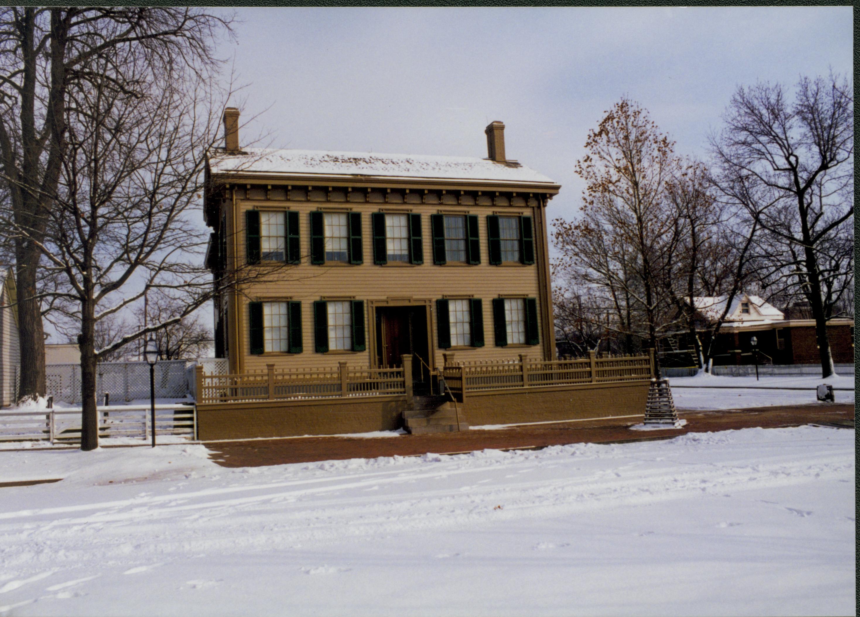 Lincoln Home in the snow, cleared brick plaza in front with elm tree in cage on right. Arnold House in background on right.  Maintenace vehicle tracks and footprints visible in snow on 8th Street. Edge of Corneau house on far left. Looking East/Southeast from boardwalk on west side of 8th Street snow, Lincoln Home, Arnold, Corneau, brick plaza, elm tree, 8th Street