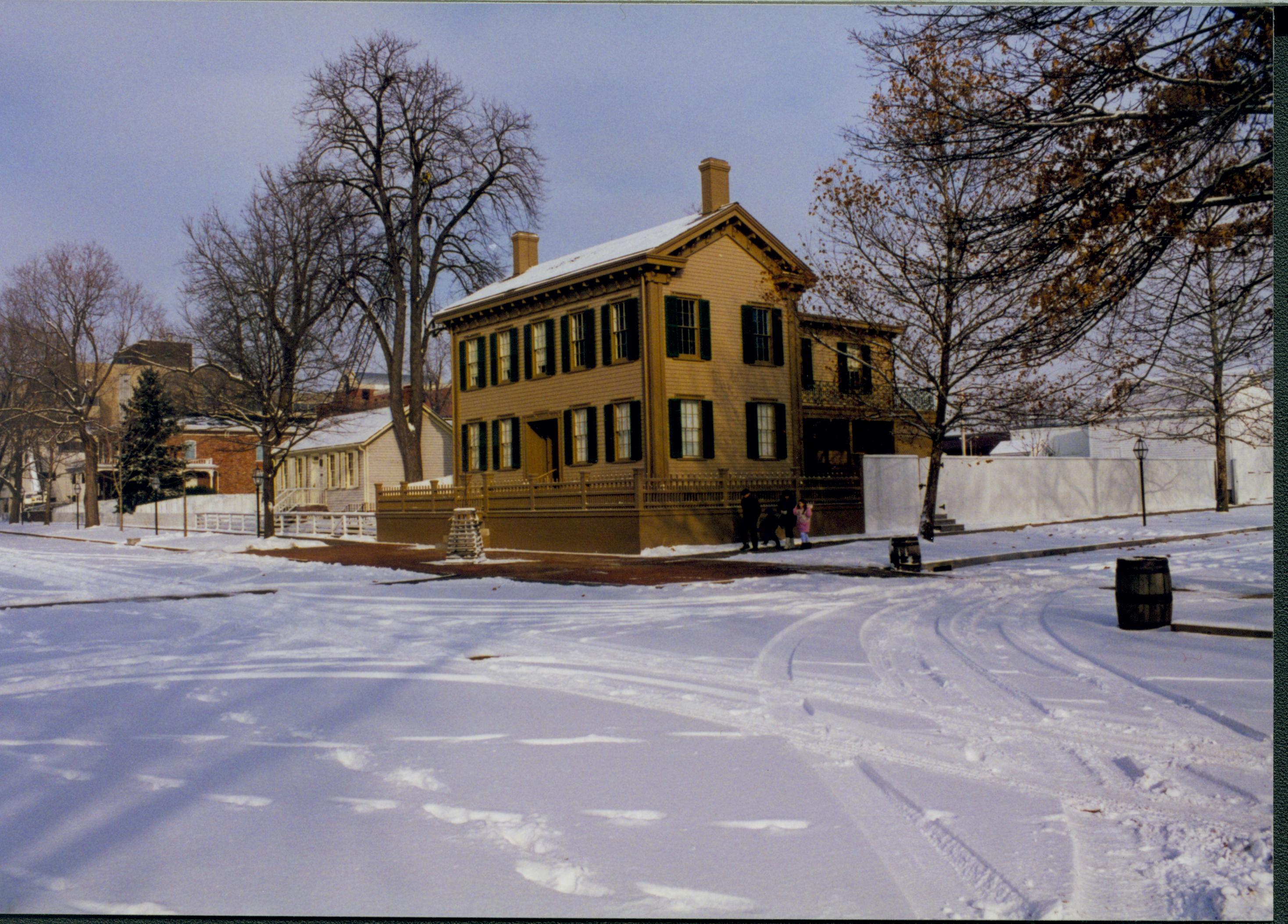 Lincoln neighborhood in snow.  Lincoln Home in center with cleared brick plaza in front and elm tree in cage on plaza. Corneau House on left, Conference Center in brick on far left.  Trash barrels on corners of 8th and Jackson Street intersection.  Maintenance vehicle tracks and footprints visible in snow. Visitors on boardwalk along Jackson Street near plaza. Looking Northeast from just south of 8th and Jackson Streets snow, Lincoln Home, Corneau, Conference Center, 8th Street, Jackson Street, brick plaza, elm tree