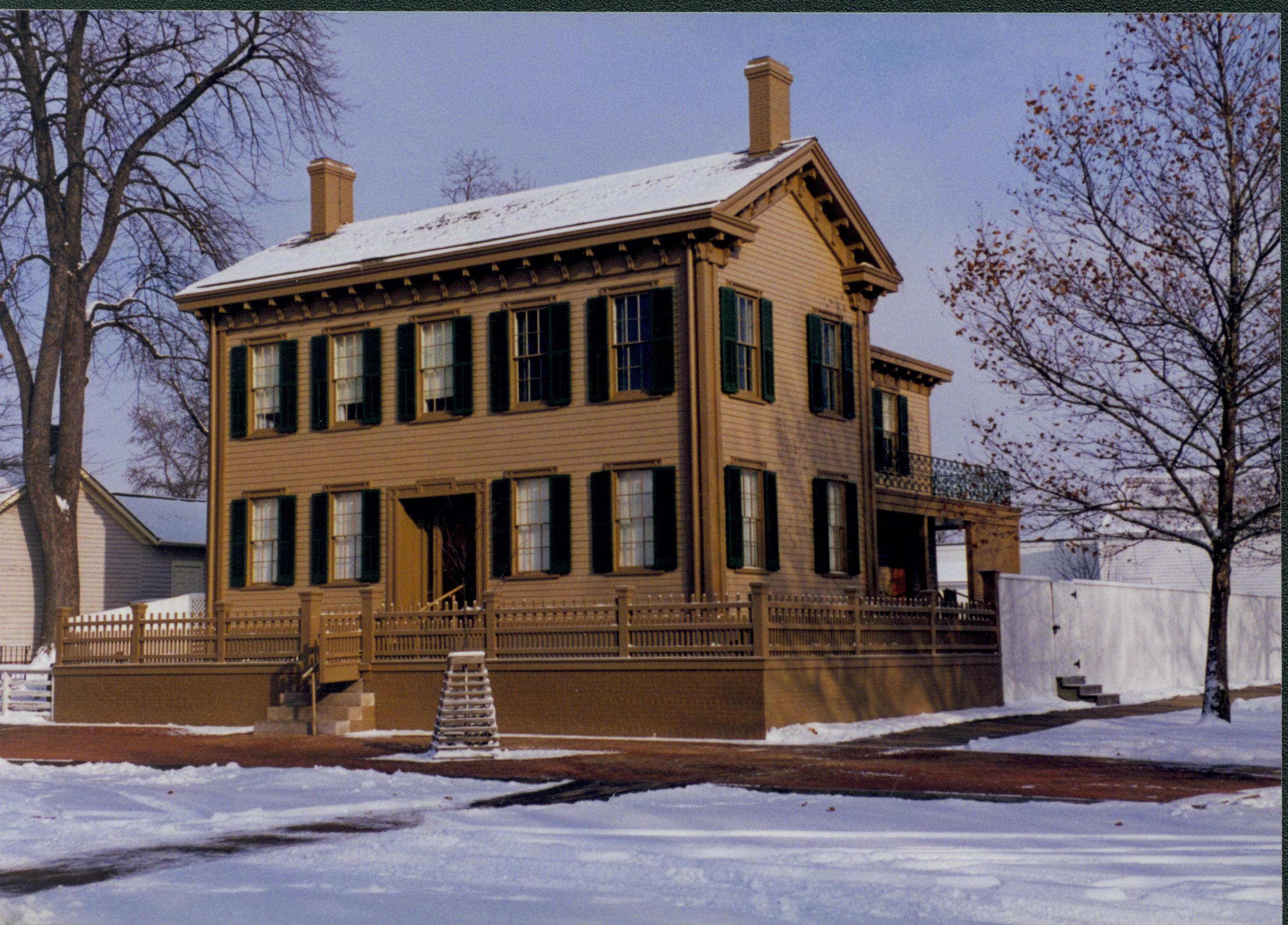 Lincoln Home in the snow with cleared brick plaza in front. Elm tree in cage on plaza. Cleared boardwalks visible on South side of house into 8th Street. Corneau House on left. Looking Northeast from 8th and Jackson Street intersection snow, Lincoln Home, brick plaza, elm tree, Corneau