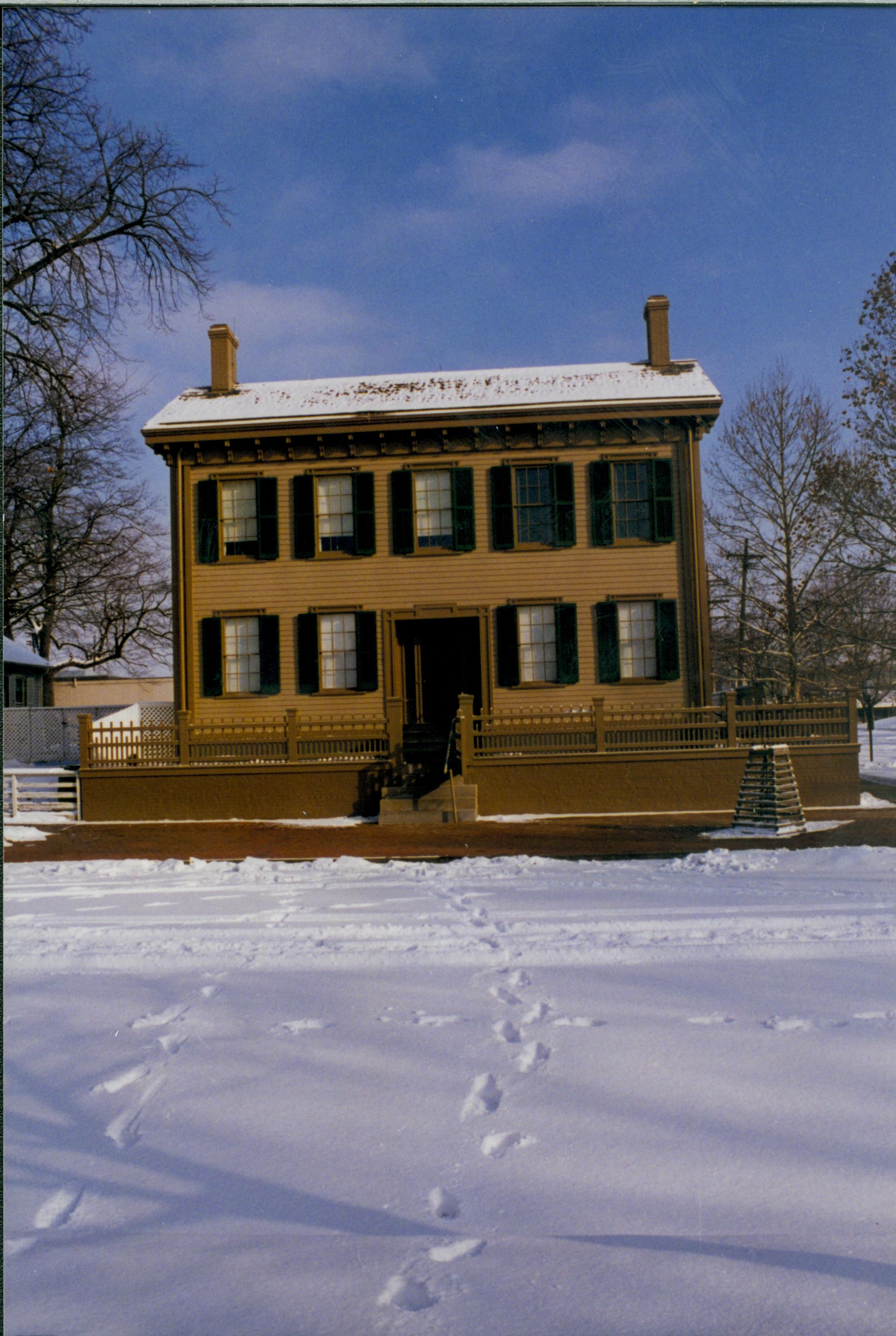 Lincoln Home in the snow with cleared brick plaza in front, elm tree in cage on right. Footprints and maintenance vehicle tracks visible in snow along 8th Street. Corneau House on far left. B-2 visible between Lincoln and Corneau houses Looking East from boardwalk on west side of 8th Street. snow, Lincoln Home, Corneau, B-2, brick plaza, elm tree