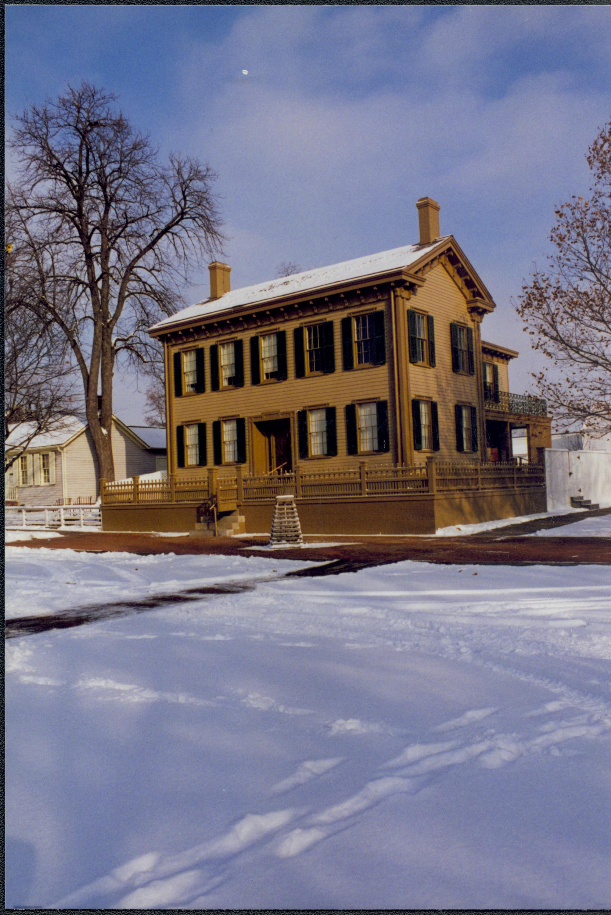 Lincoln Home in the snow with cleared brick plaza in front and elm tree in cage on plaza. Corneau House on left. Footprints and maintenance vehicle tracks visible in snow along 8th and Jackson Street.  Looking Northeast from 8th and Jackson Street intersection snow, Lincoln Home, Corneau, 8th Street, Jackson Street, brick plaza, elm tree