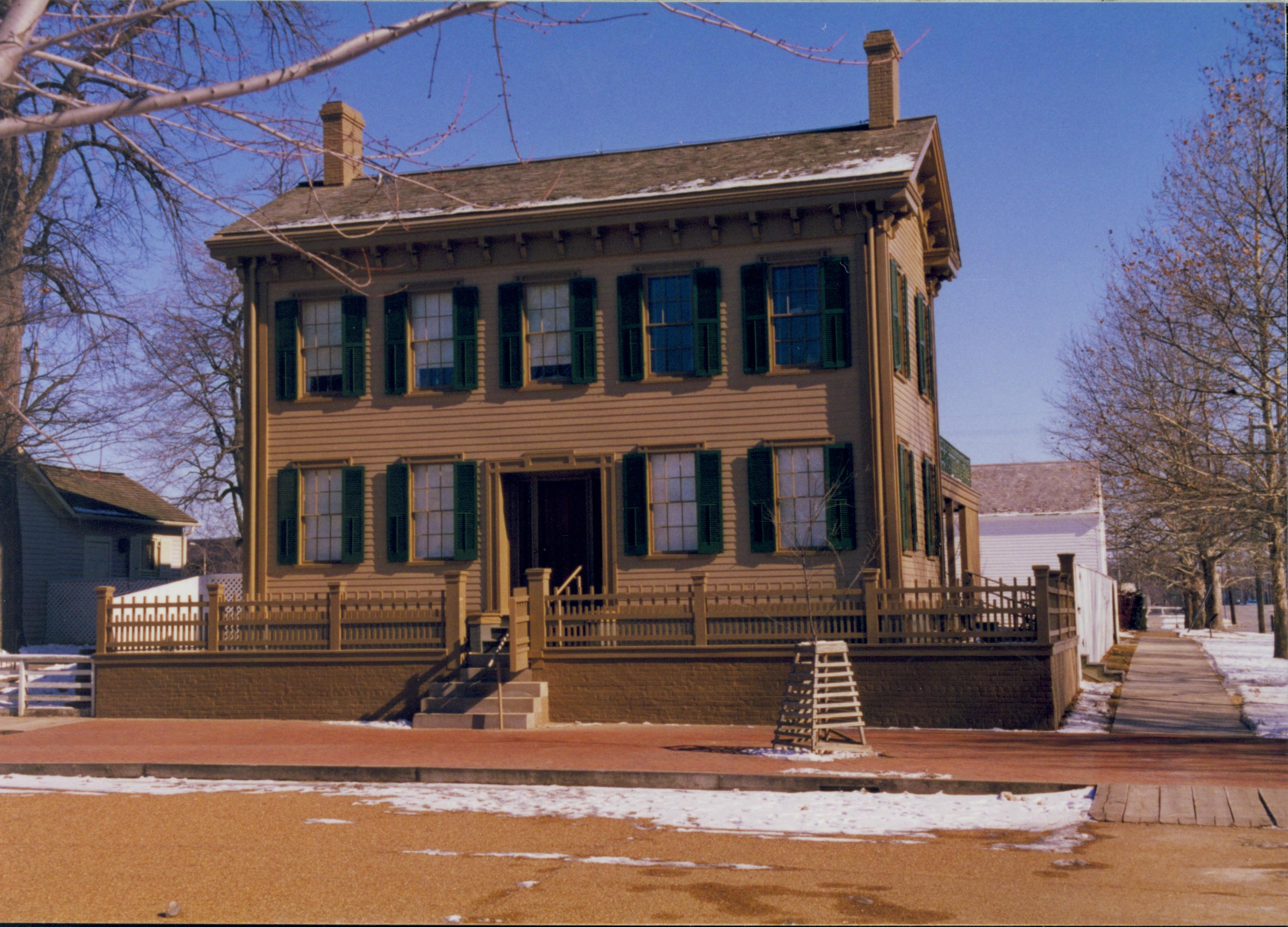 Lincoln Home in winter with scattering of snow. Cleared brick plaze in front with elm tree in cage on right.  Cleared boardwalk on right. Lincoln Barn in background on right.  Corneau House on left. Looking East/Northeast from 8th and Jackson Street intersection snow, Lincoln Home, Lincoln Barn, boardwalk, brick plaza, elm tree, Corneau, 8th Street, Jackson Street