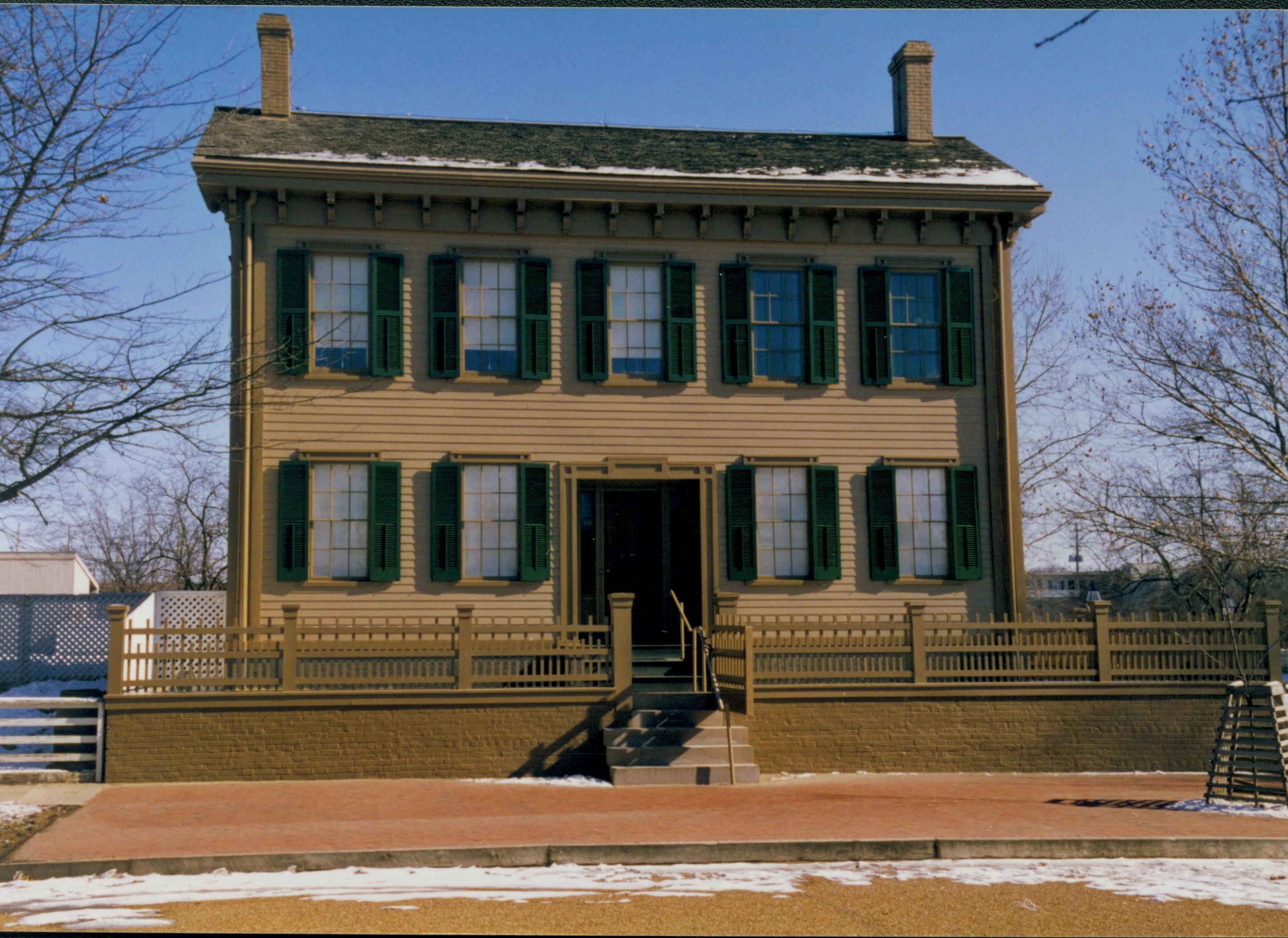 Lincoln Home in winter with some snow. Clear brick plaze in front, elm tree in cage on right.  Fence for Carrigan lot on left with B-2 in background. Looking East from 8th Street snow, Lincoln Home, B-2, brick plaza, elm tree